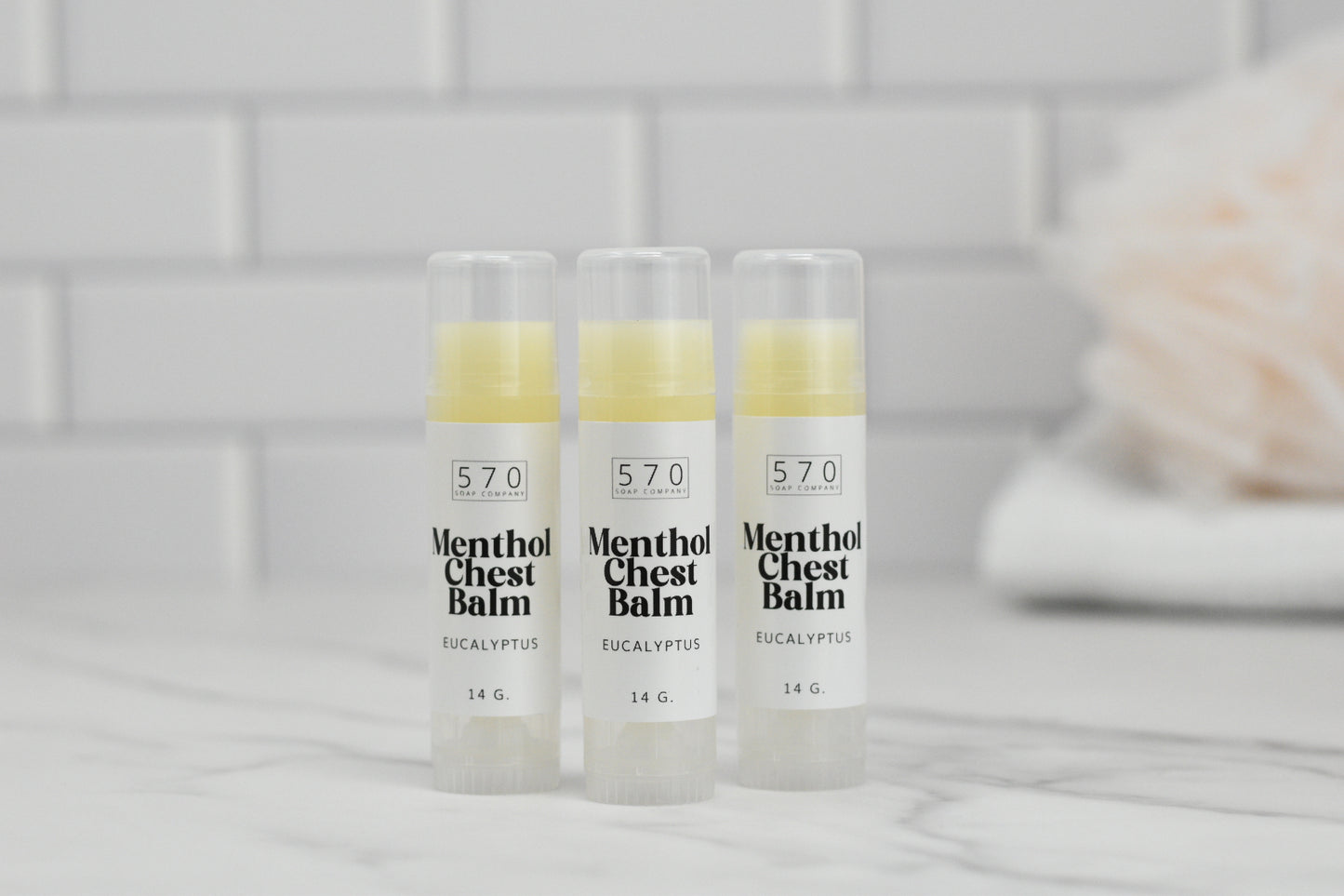 Three tubes of 570 Soap Co.'s Menthol Chest Balm - Eucalyptus are placed on a marble surface against a backdrop of white tiles. Each 14-gram tube, packed with natural ingredients, is standing upright. In the background, a blurred towel hints at relief from respiratory discomfort.