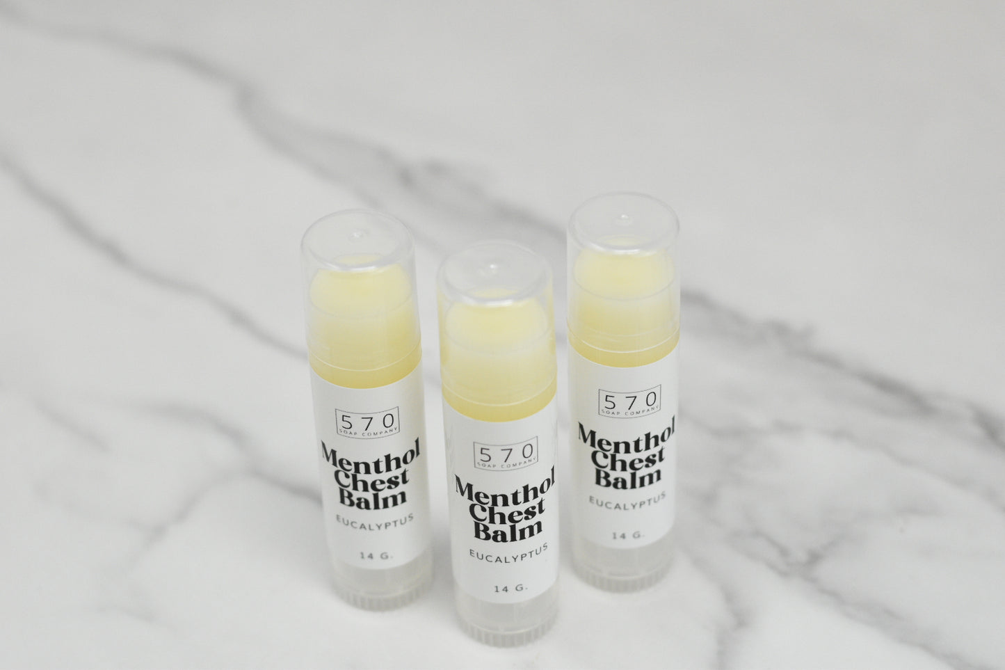 Three tubes of Menthol Chest Balm - Eucalyptus from 570 Soap Co. stand upright on a marble surface. Each 14g tube is crafted to ease respiratory discomfort and features a blend of natural ingredients. Their clear caps add an elegant touch to their healing promise.