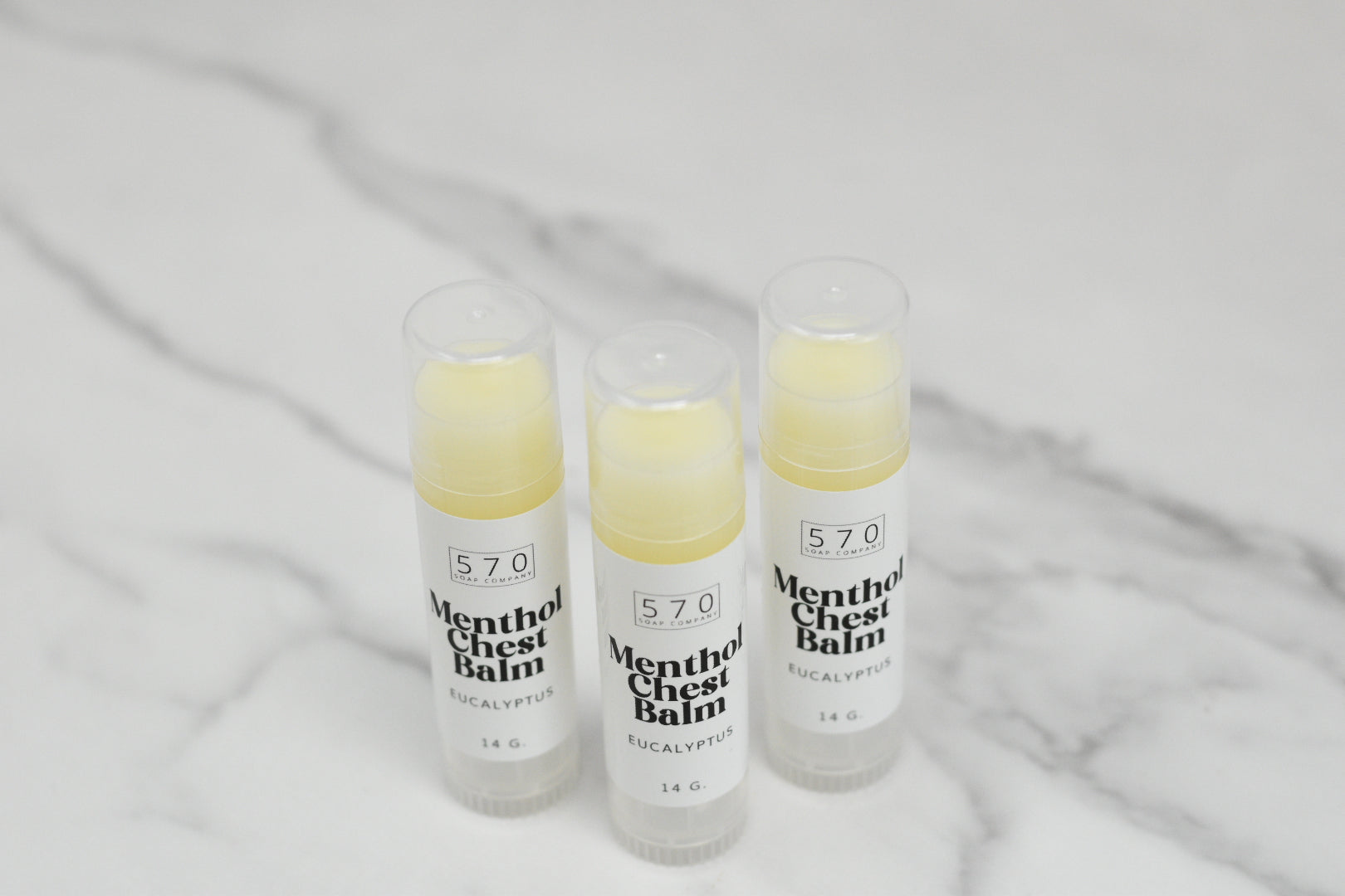 Three tubes of Menthol Chest Balm - Eucalyptus from 570 Soap Co. stand upright on a marble surface. Each 14g tube is crafted to ease respiratory discomfort and features a blend of natural ingredients. Their clear caps add an elegant touch to their healing promise.