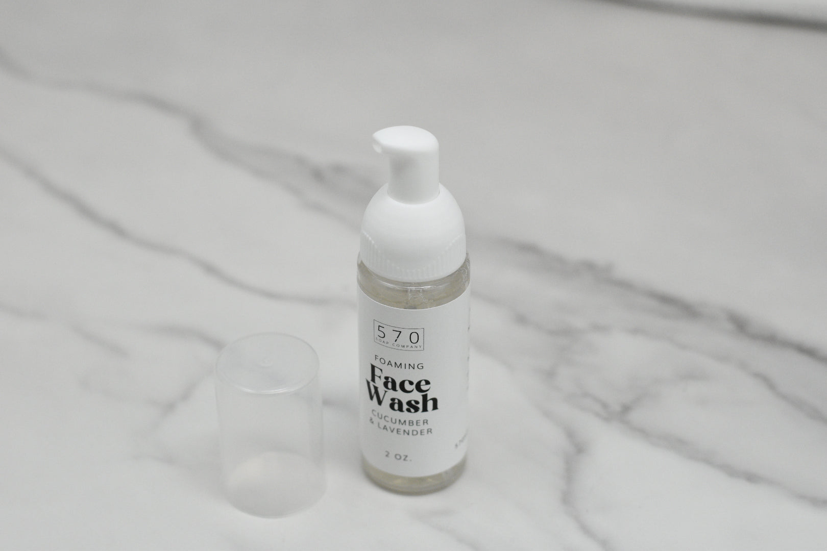 A 2 oz bottle of 570 Soap Co.'s Foaming Face Wash - Cucumber Lavender, enhanced with Lavender Floral Water, sits gracefully on a white marble surface. Its pump dispenser and soothing cucumber lavender fragrance make it a refreshing choice. The cap lies beside the container, completing this serene scene.