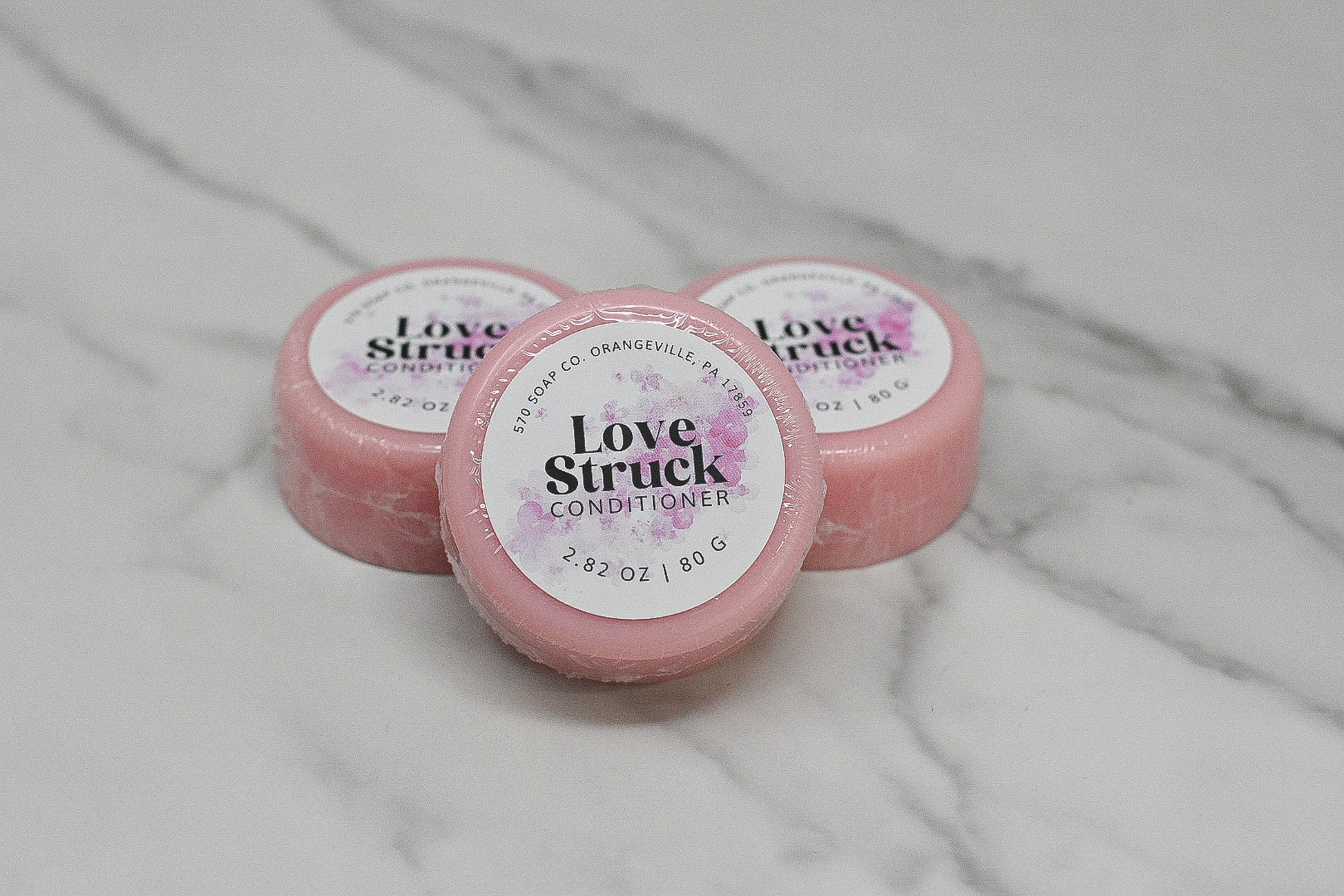 Three small pink containers of the Conditioner Bar for All Hair Types - Love Struck by 570 Soap Co. are displayed on a marble surface. Each eco-friendly option is marked with the brand name and weight (2.8 oz/80 g), showcasing a luxurious floral design for an elevated hair care experience.