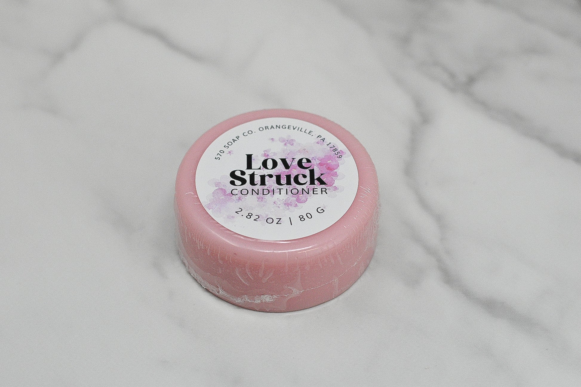 The Love Struck Conditioner Bar for All Hair Types by 570 Soap Co. in Orangeville, PA provides a luxurious hair care experience. Weighing 2.82 oz (80 g), this eco-friendly alternative comes in a pink round container adorned with a purple floral design, elegantly displayed on a white marble surface.