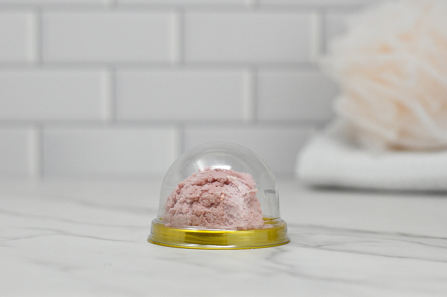 A Solid Bubble Bath Scoop from 570 Soap Co. is elegantly presented under a clear plastic dome with a gold base on a marble counter, resembling a scoop of pink ice cream. The background showcases a blurred view of a white tiled wall and plush item, capturing the charm of luxurious bubbles.