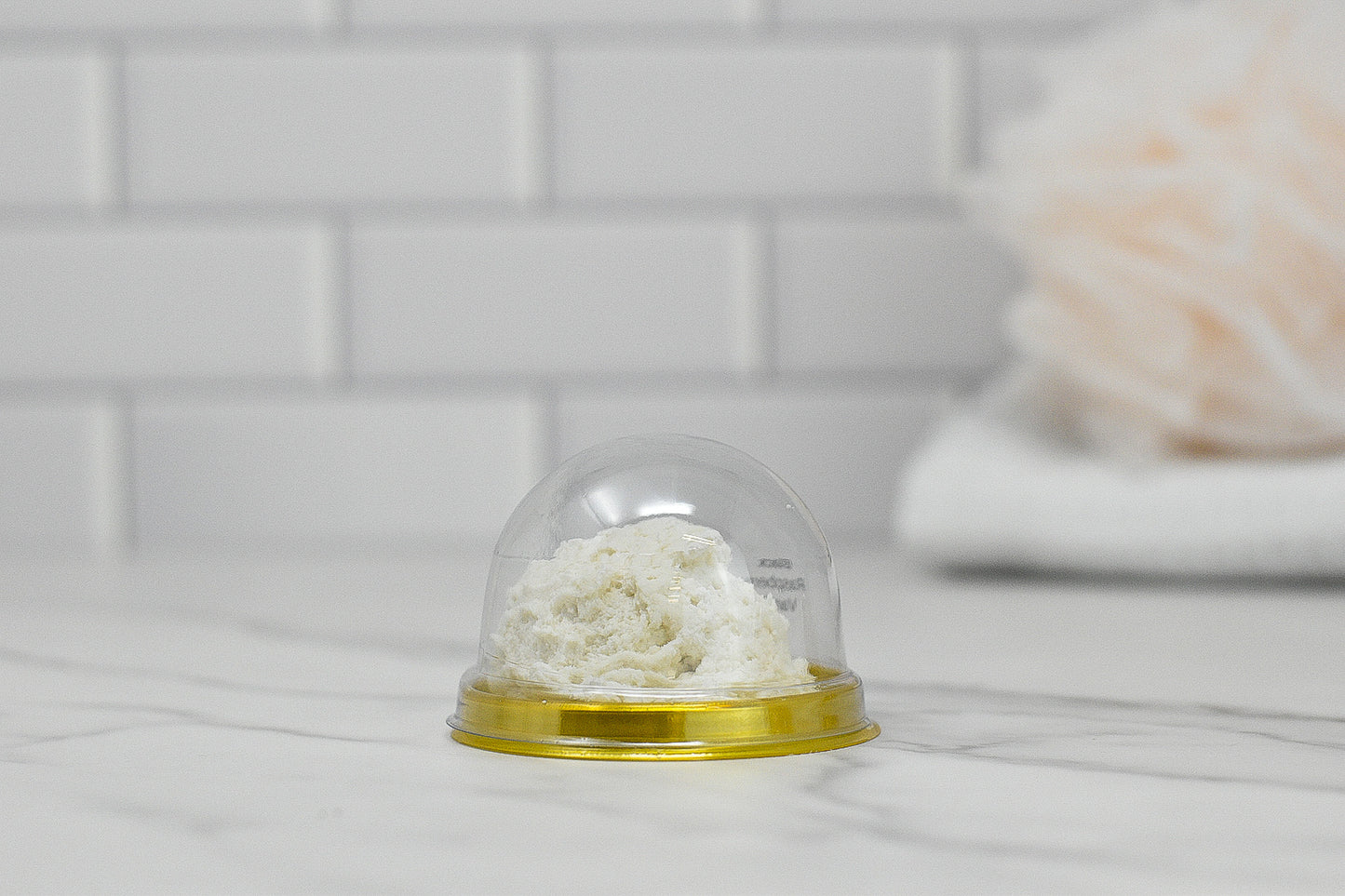 A small, clear plastic dome container with a yellow base contains a portion of white fluffy substance that resembles the Solid Bubble Bath Scoop from 570 Soap Co. It rests on a marble counter amidst luxurious bubbles, with a backdrop of white subway tiles and a blurred light pink decorative object.