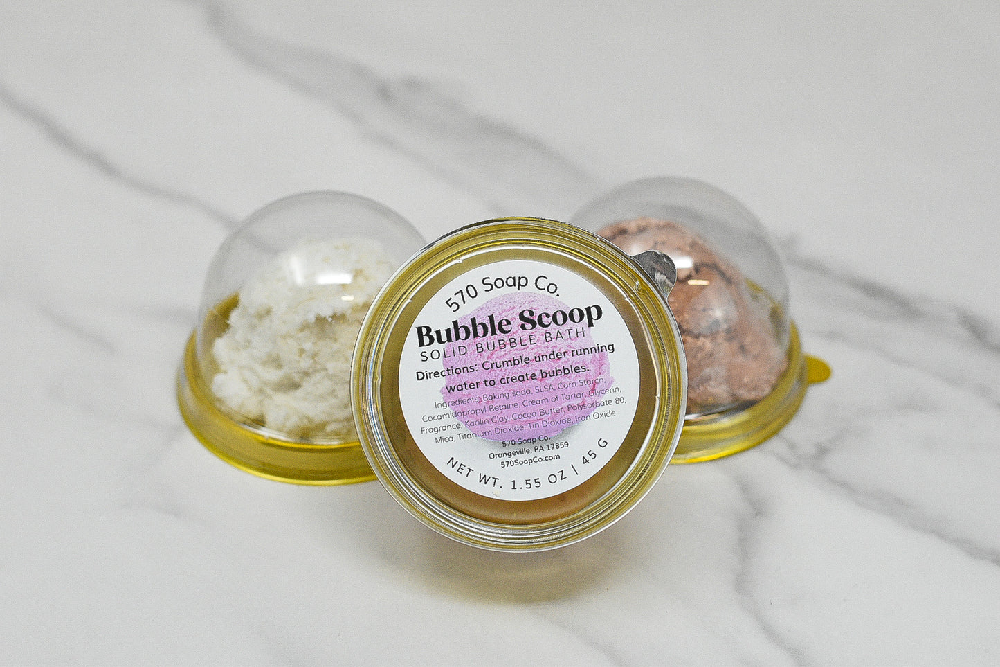 Three dome-shaped Solid Bubble Bath Scoops from 570 Soap Co. are elegantly arranged on a marble surface. The center scoop, clearly labeled Solid Bubble Bath Scoop, is in focus, highlighting its skin-caring formula directions and weight details, while the luxurious bubbles promise a pampering experience. The other scoops blur softly in the background.