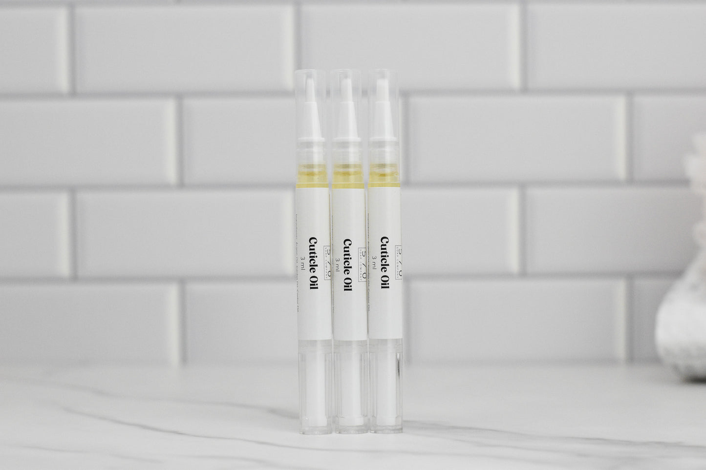 Three pen-style applicators of 570 Soap Co.'s Nourishing Cuticle Oil, enriched with argan oil, are displayed upright on a white marble surface. The background showcases a sleek white subway tile wall.