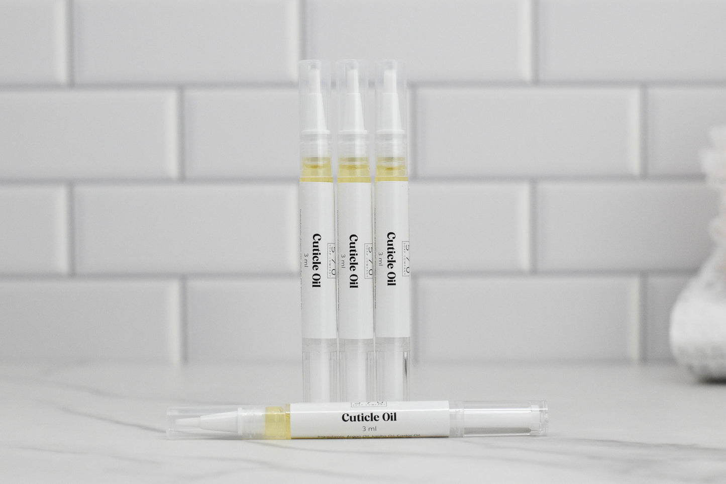 Clear pen-style tubes of 570 Soap Co.'s Nourishing Cuticle Oil are arranged on a marble surface, enriched with Vitamin E. One tube rests horizontally while three stand upright against a backdrop of white subway tiles.