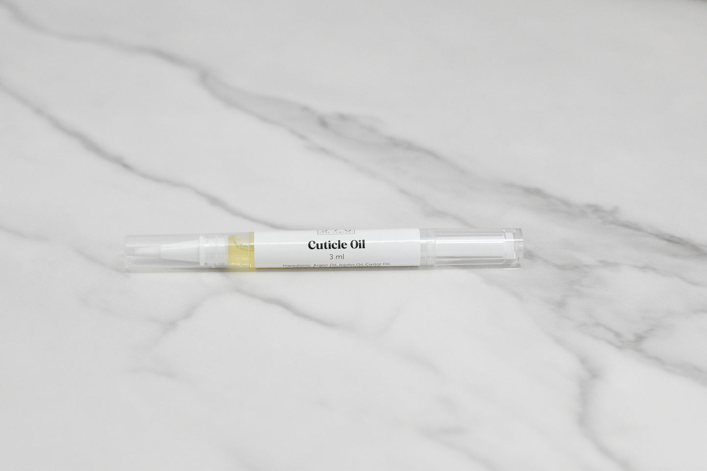 A pen-shaped container of Nourishing Cuticle Oil from 570 Soap Co., labeled 3 in 1, sits on a white marble surface. The transparent casing reveals a light yellow liquid enriched with Vitamin E, designed to revitalize and protect.