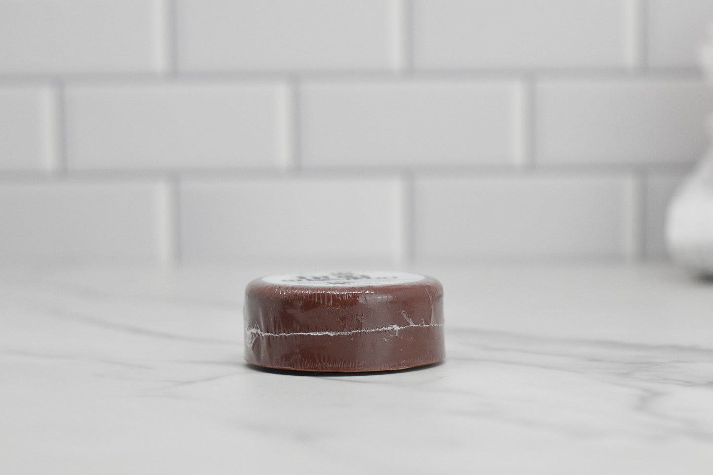 A small, round bar of 570 Soap Co.'s Dog Shampoo Bar - All Natural with a soothing lavender scent sits on a white marble countertop against a backdrop of white subway tiles. Its brown packaging features visible seam lines, providing a gentle cleansing sensation to your senses even before it's unwrapped.