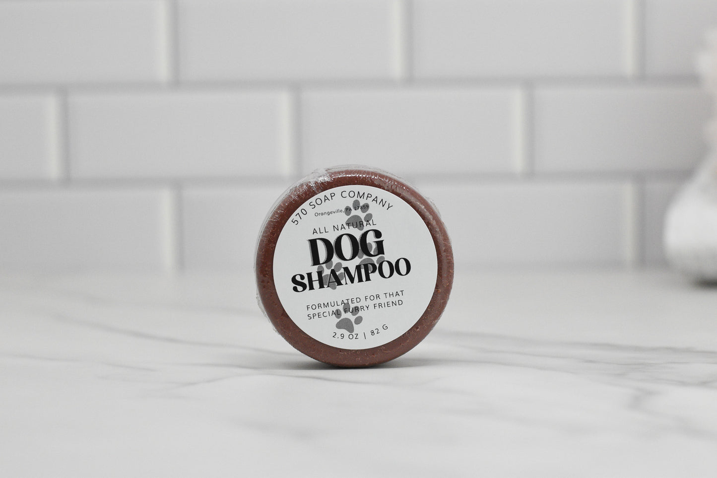 A small, round container of 570 Soap Co.'s Dog Shampoo Bar - All Natural with a soothing lavender scent rests on a marble surface. The label reads, "Formulated for that Special Furry Friend," and it weighs 2.9 oz (81.6 g). A tiled wall serves as the background, highlighting the promise of gentle cleansing for your pet's comfort.