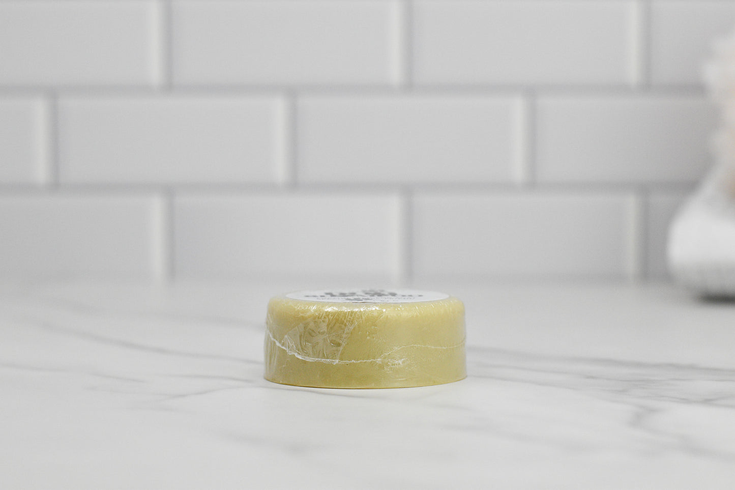 A round, beige bar of Dog Shampoo Bar - All Natural by 570 Soap Co., encased in clear plastic, rests on a marble countertop. Behind it, a white subway tile wall forms a spotless, minimalist backdrop. This gentle soap infuses your pet's bath time with the relaxing aroma of lavender.