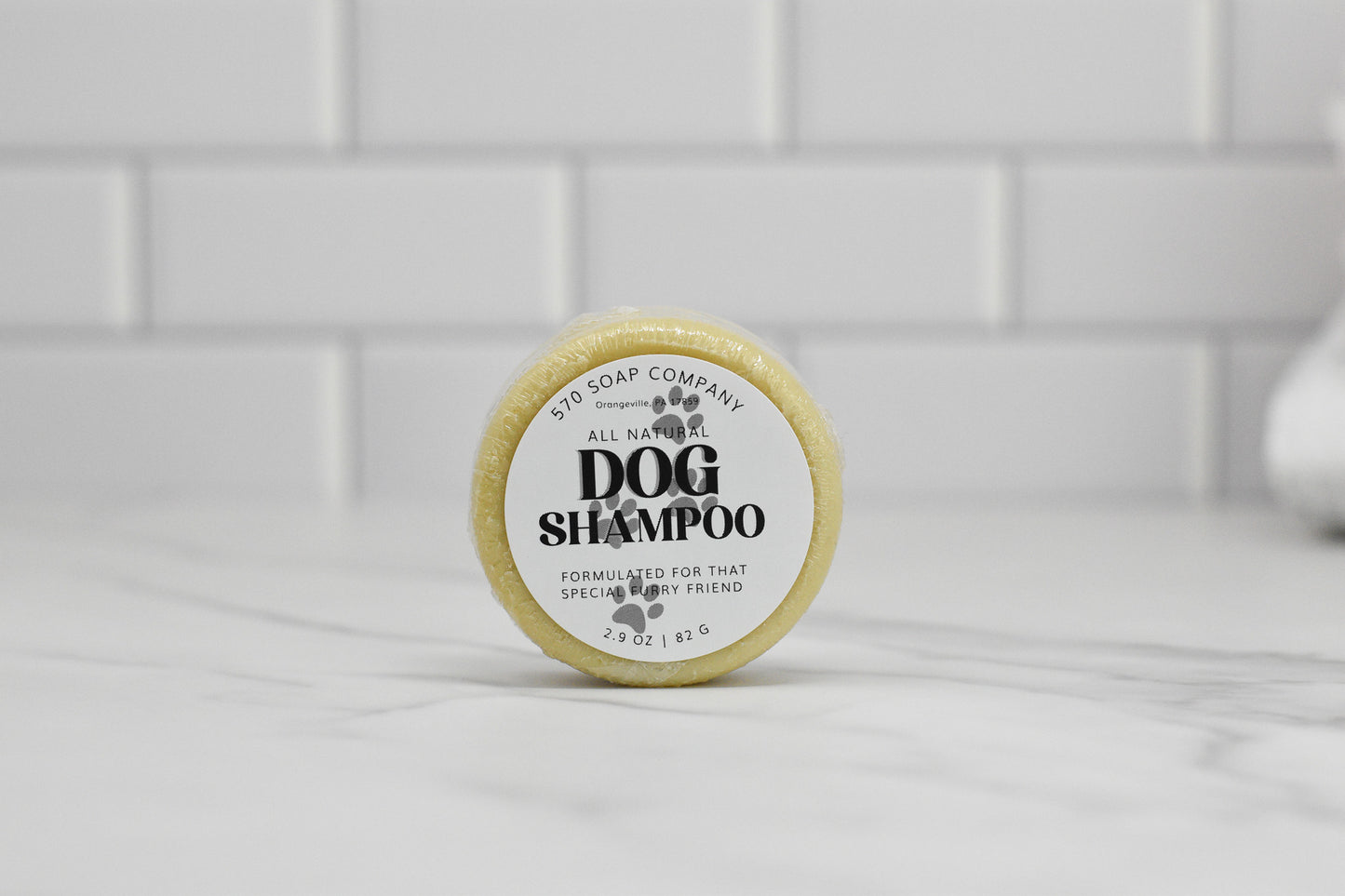 A round dog shampoo bar made with all-natural ingredients and featuring a calming lavender scent is displayed on a marble surface. The label reads 570 Soap Co. Dog Shampoo Bar - All Natural, specially formulated for that special furry friend. A subway tile backsplash in the background accentuates its promise of gentle cleansing.