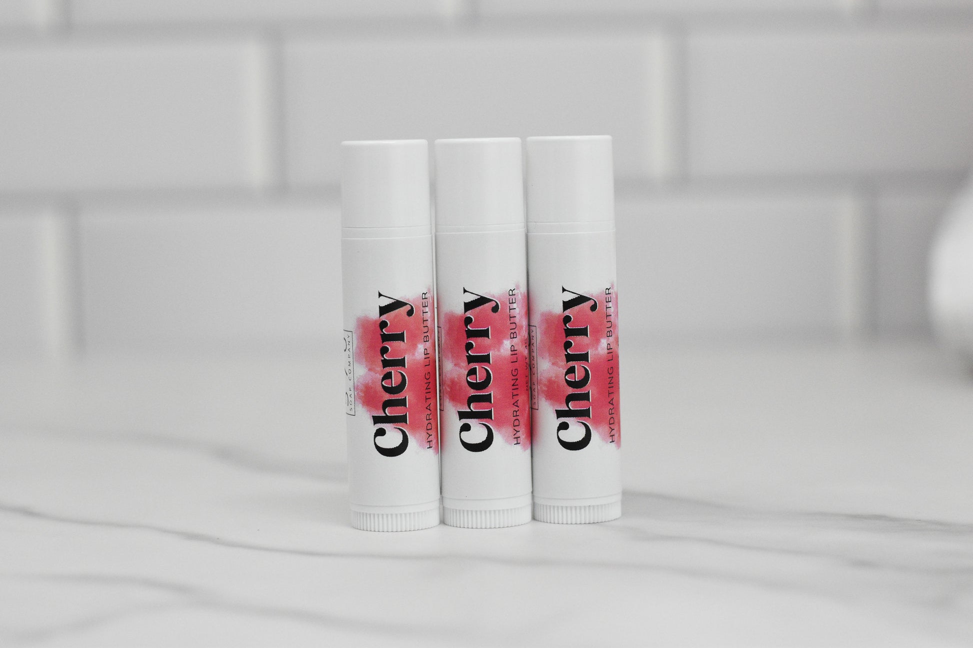Three tubes of Cherry Hydrating Lip Balm from 570 Soap Co., featuring natural ingredients, stand upright on a marble surface. The white tubes showcase the word "Cherry" in bold black letters highlighted with a red accent. Enriched with nourishing oils, they are placed against a tiled white background.