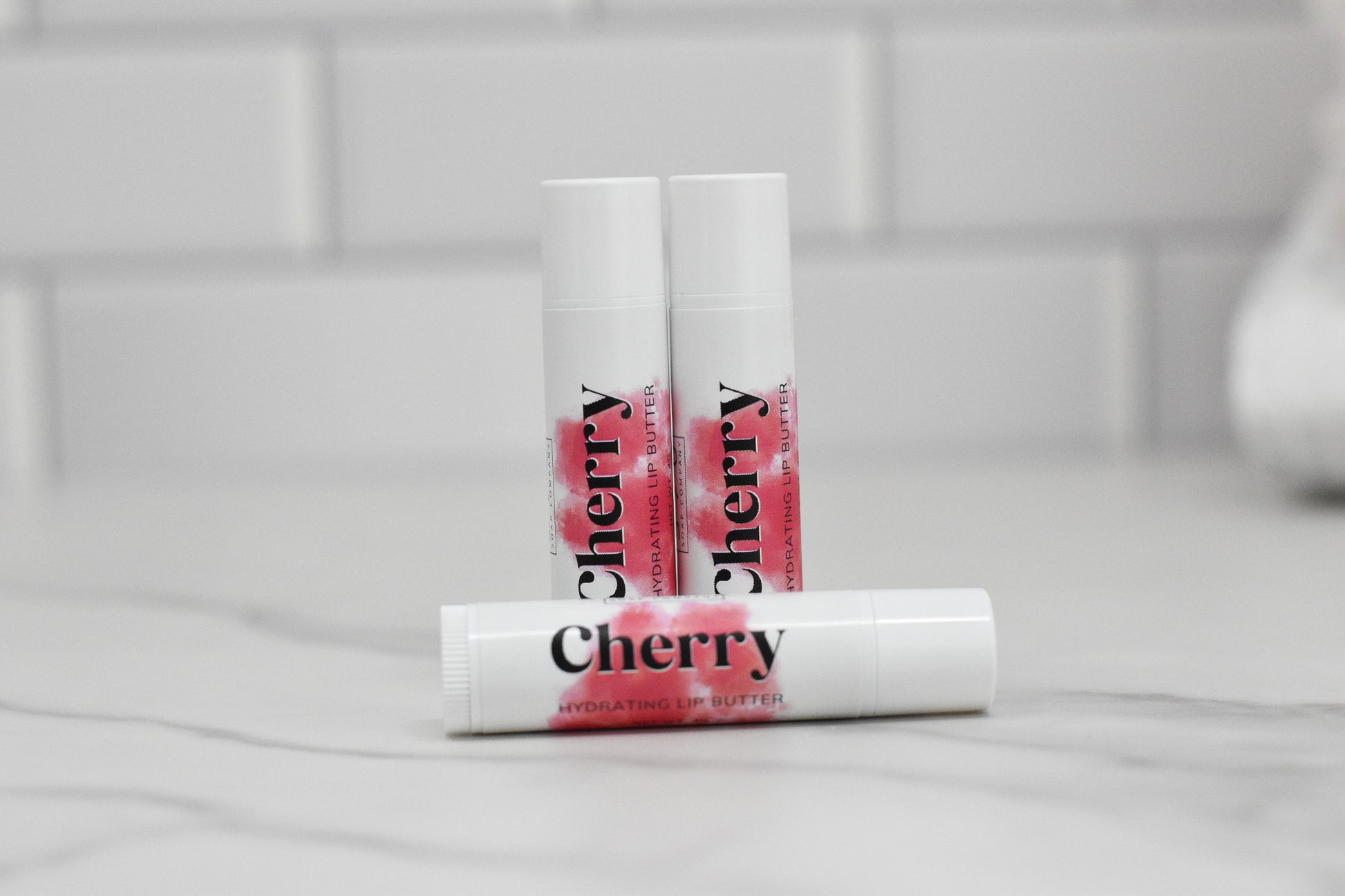 Three white tubes of Cherry Hydrating Lip Balm from 570 Soap Co. are displayed. Two stand upright in the background, while one lies horizontally in the foreground on a gray marbled surface. Infused with nourishing oils, these luxurious balms rest against a tiled wall, promising intense hydration.