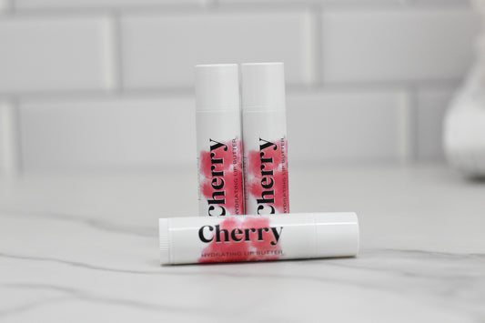 Three white tubes of Cherry Hydrating Lip Balm from 570 Soap Co. are displayed. Two stand upright in the background, while one lies horizontally in the foreground on a gray marbled surface. Infused with nourishing oils, these luxurious balms rest against a tiled wall, promising intense hydration.