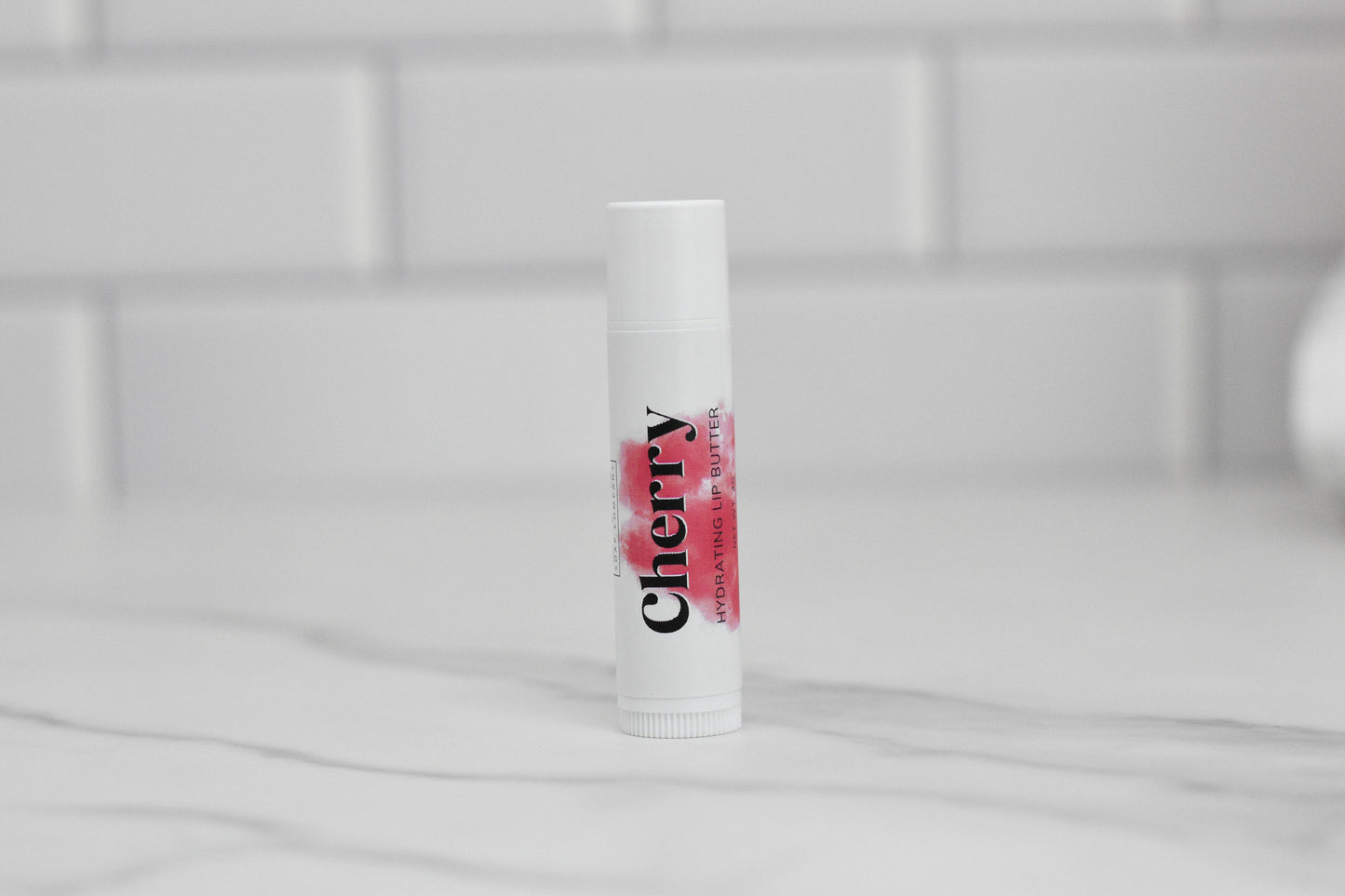 A tube of Cherry Hydrating Lip Balm by 570 Soap Co., enriched with nourishing oils and natural ingredients, stands upright on a marble countertop with a blurred white tiled wall in the background.