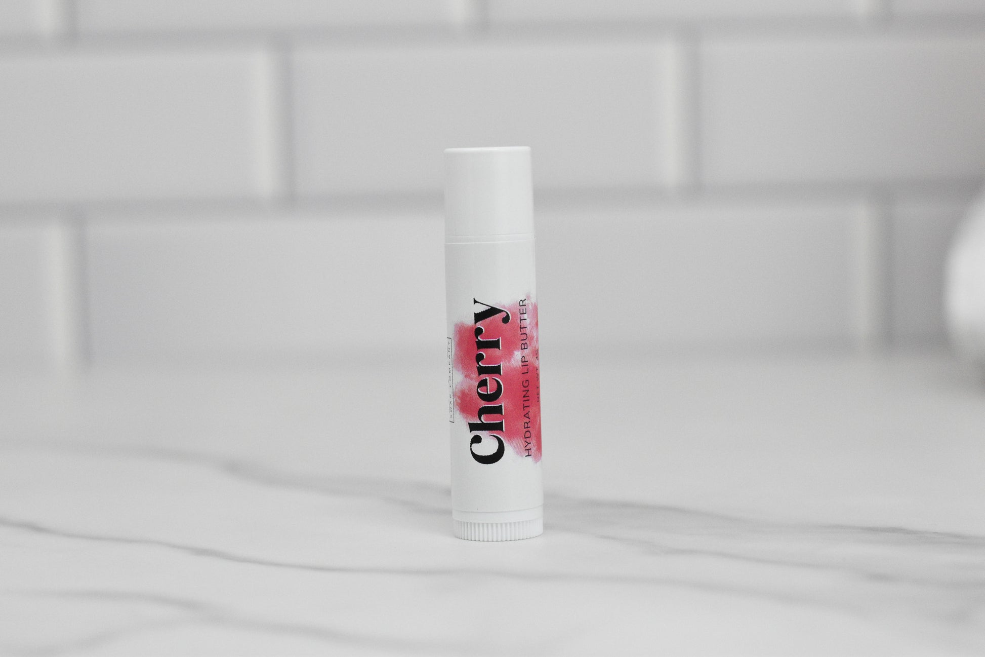 A tube of Cherry Hydrating Lip Balm by 570 Soap Co., enriched with nourishing oils and natural ingredients, stands upright on a marble countertop with a blurred white tiled wall in the background.