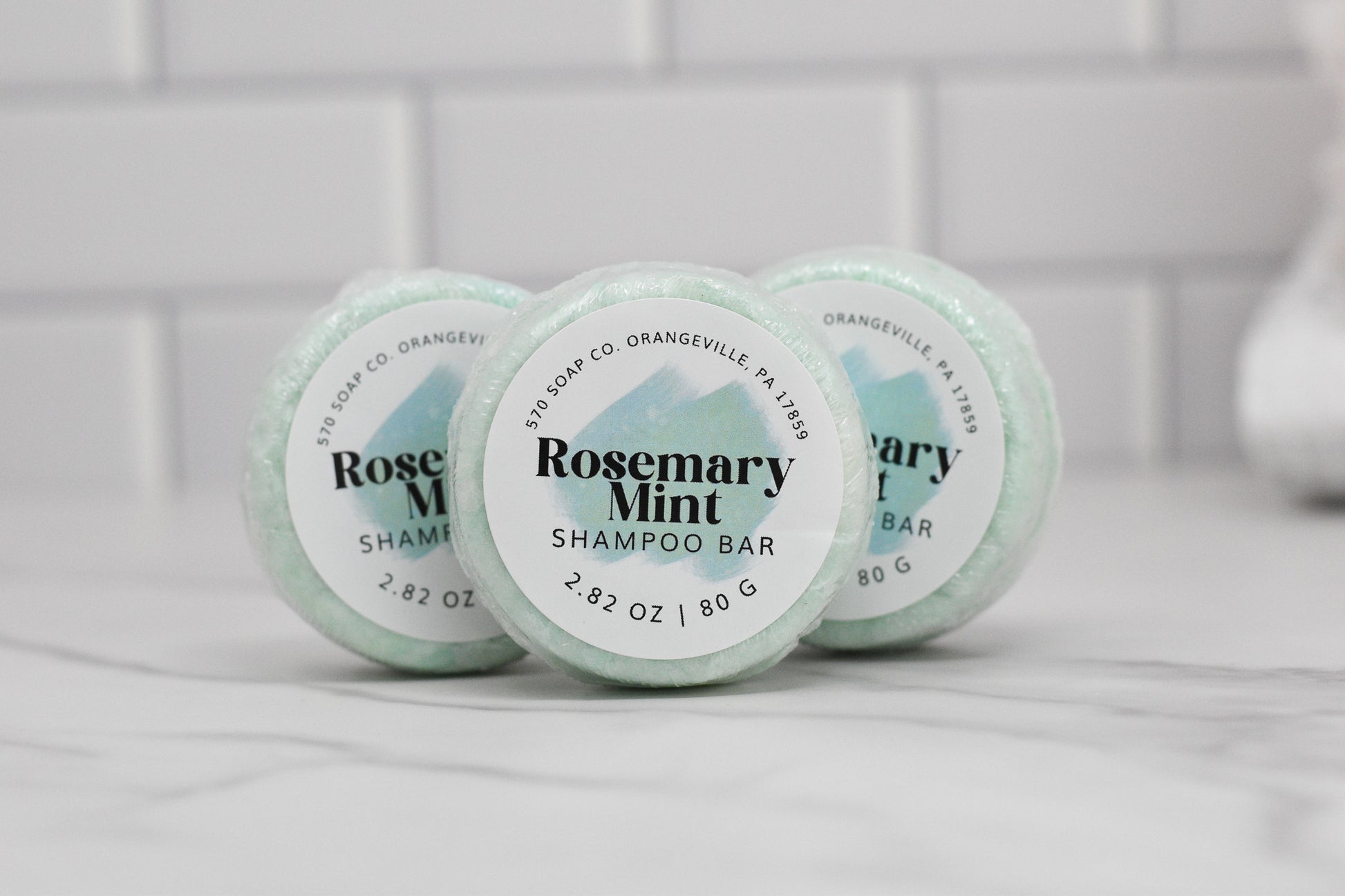 A trio of round Rosemary Mint Essential Oil Shampoo Bars from 570 Soap Co. is displayed on a white marble surface. The packaging features a light green color with white and black text. A tiled wall in the background accentuates the invigorating feel of peppermint essential oil.