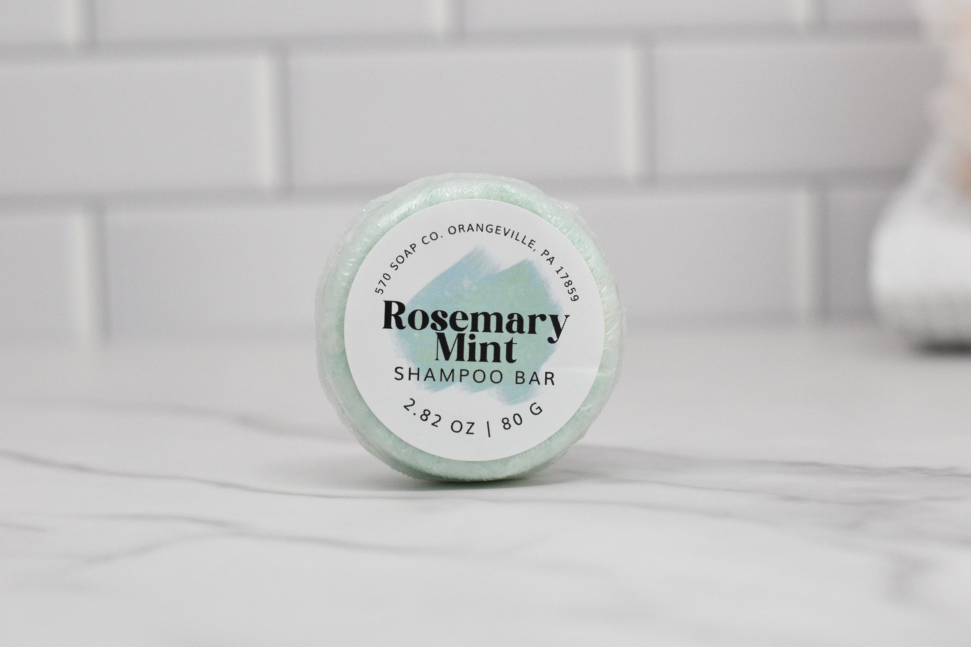 A round, pale green Rosemary Mint Essential Oil Shampoo Bar imbued with rosemary and peppermint essential oils rests on a white marble surface. The packaging reveals it weighs 2.82 oz (80g), crafted by 570 Soap Co. in Orangeville, PA. White subway tiles form a fresh and clean backdrop.