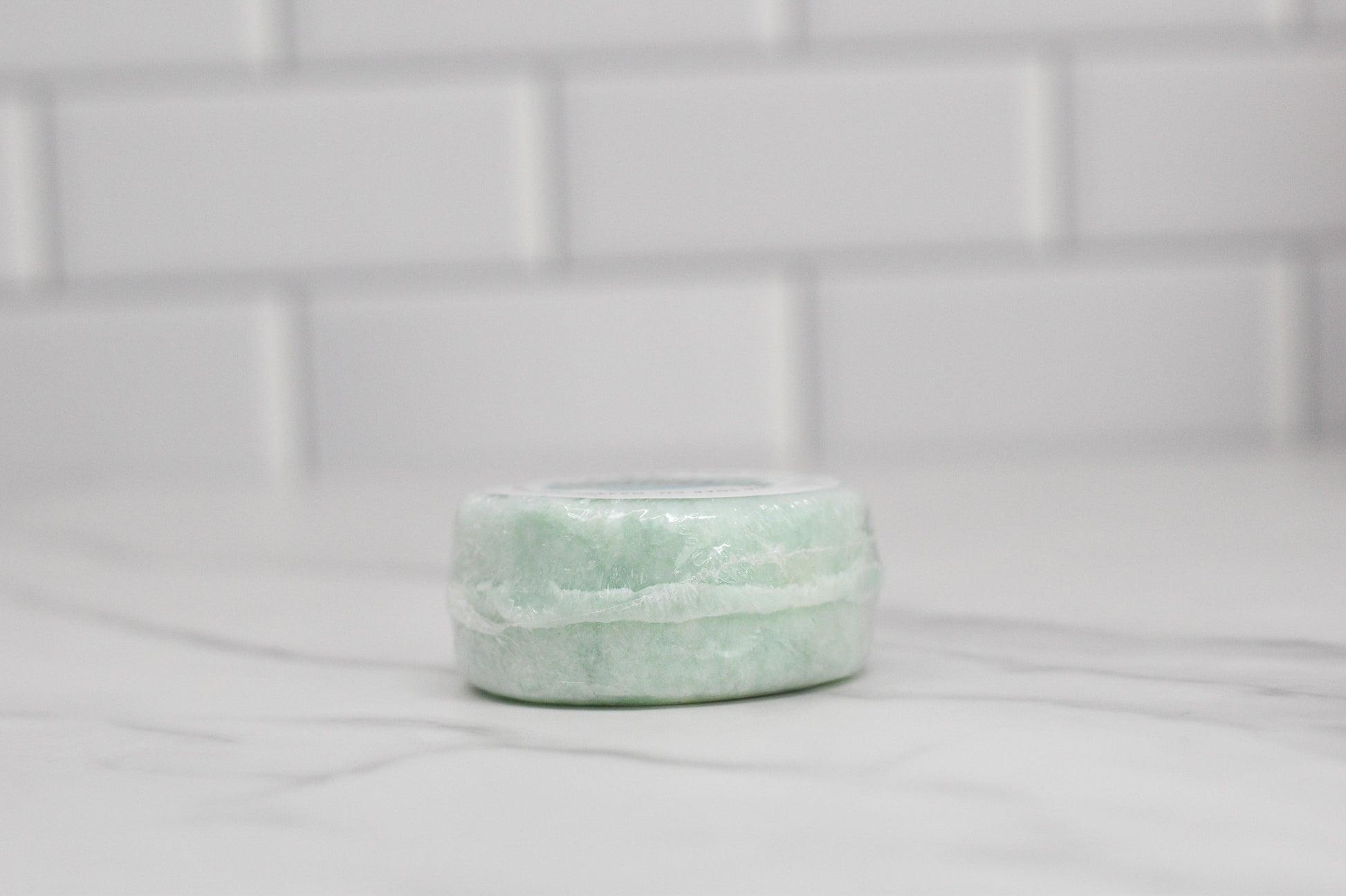 A circular, slightly translucent light green Rosemary Mint Essential Oil Shampoo Bar by 570 Soap Co. is displayed on a white marble surface with a white tiled background.