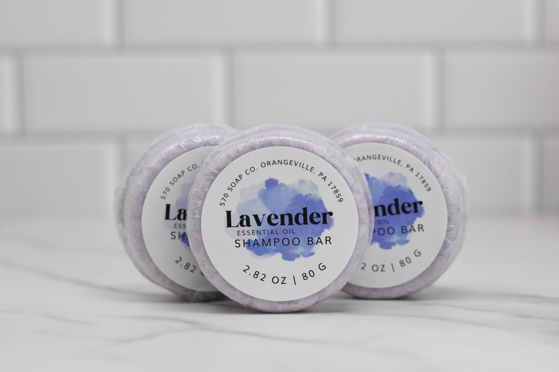Three Lavender Essential Oil Shampoo Bars, produced by 570 Soap Co., are stacked against a white tile background, making them perfect for any haircare routine. Each bar is clearly labeled with its weight, 2.82 oz / 80 g, and originates from Orangeville, PA 17859.