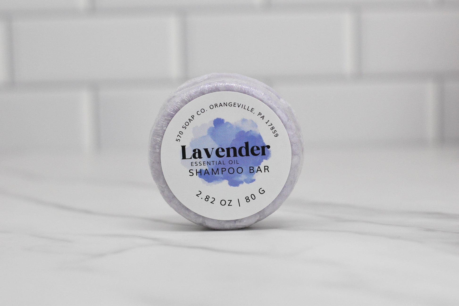 A 2.82 oz (80 g) Lavender Essential Oil Shampoo Bar from 570 Soap Co. lies on a marble surface, offering an upgrade to your haircare routine. The packaging features a watercolor lavender design set against a backdrop of white tiled walls, representing the craftsmanship of 570 Soap Co., Orangeville PA 17859.