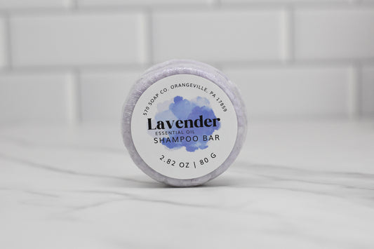 A 2.82 oz (80 g) Lavender Essential Oil Shampoo Bar from 570 Soap Co. lies on a marble surface, offering an upgrade to your haircare routine. The packaging features a watercolor lavender design set against a backdrop of white tiled walls, representing the craftsmanship of 570 Soap Co., Orangeville PA 17859.