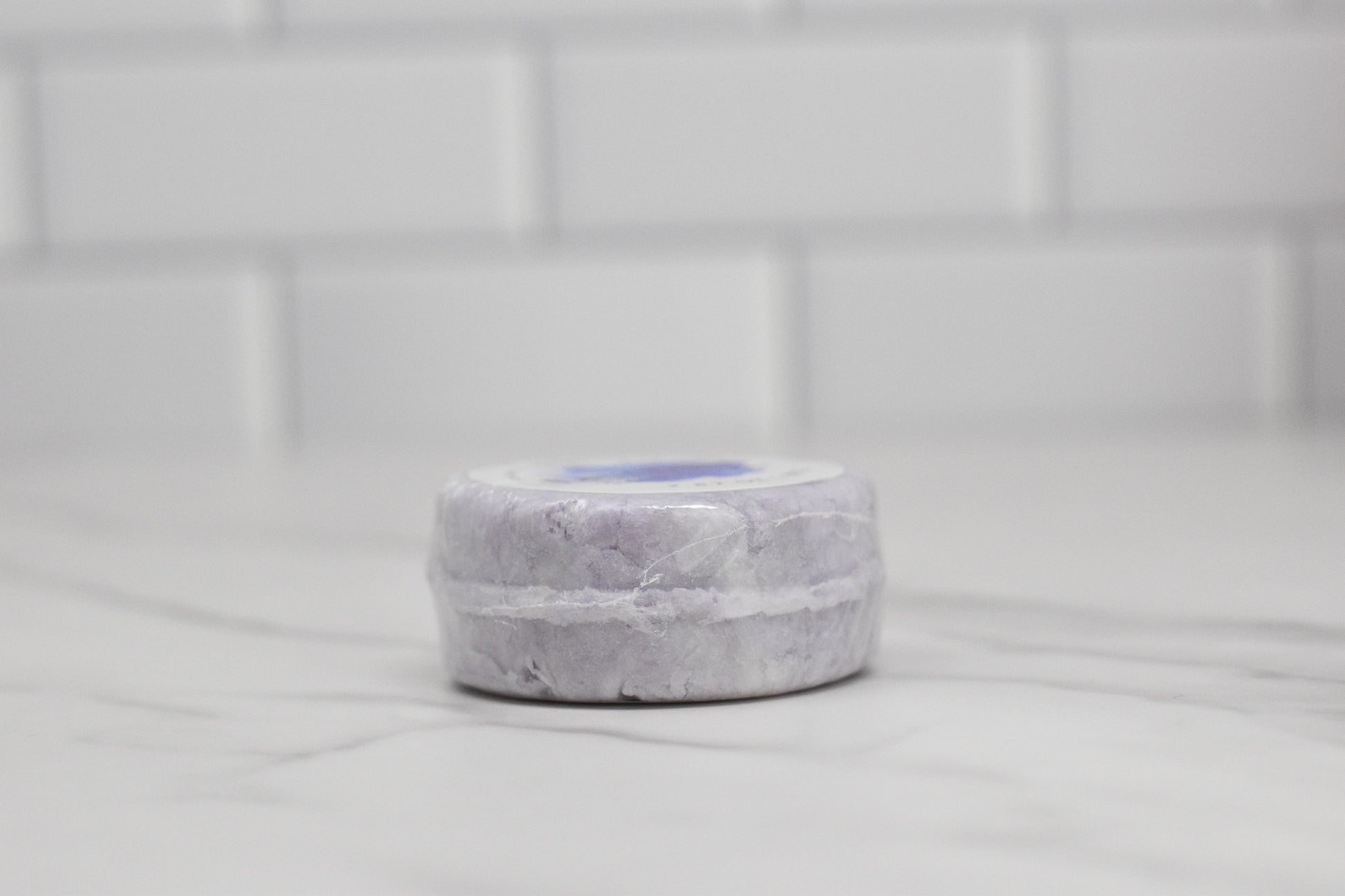 The Lavender Essential Oil Shampoo Bar from 570 Soap Co., a round lavender-colored gem, rests elegantly on a marble countertop, with a backdrop of white tiled walls that enhance its calming ambiance.