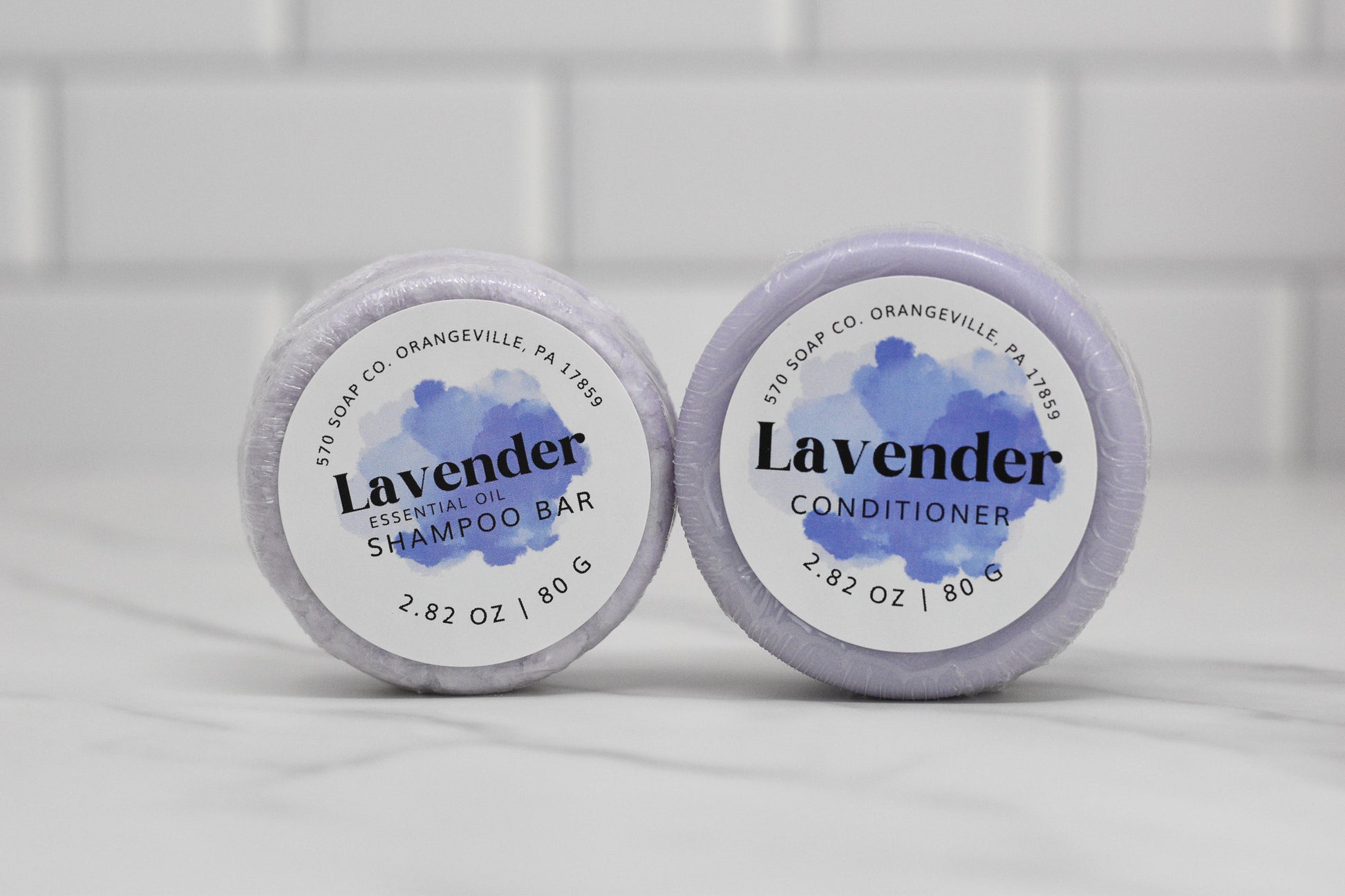 Two round bars are placed on a white surface. On the left is the Lavender Essential Oil Shampoo Bar by 570 Soap Co., while on the right is the Lavender Conditioner. They promise a soothing haircare routine, with each bar weighing 2.82 oz or 80 g, set against a tiled background.