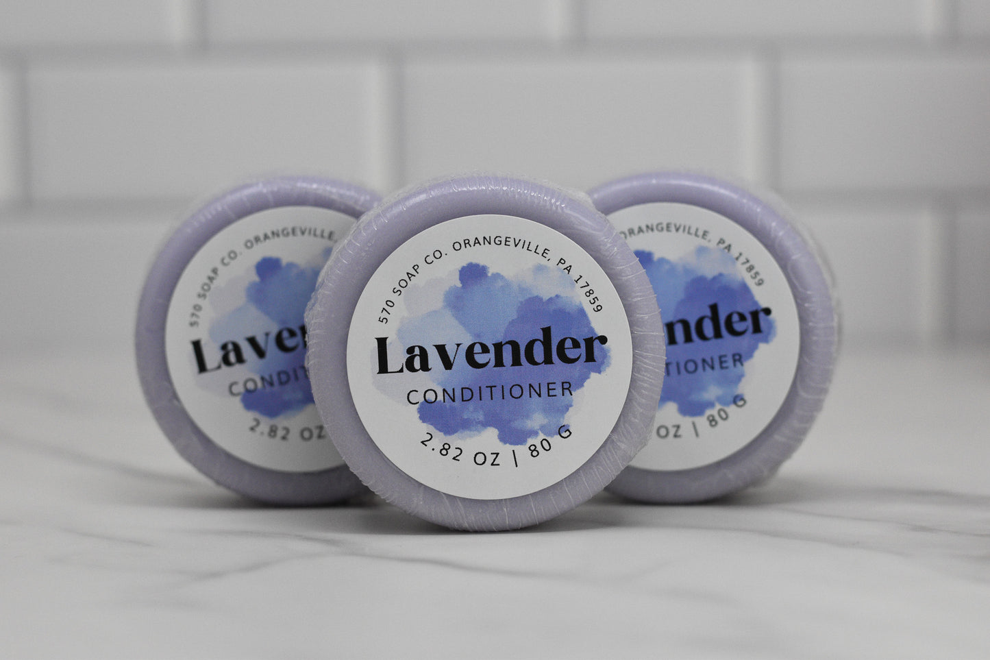 Stacked on a white tile surface are three round bars of lavender conditioner, recognized for their eco-friendly hair care benefits. Each bar is wrapped in light purple packaging, displaying the labels 570 Soap Co. Orangeville, PA 17859 and 2.82 oz / 80 g.