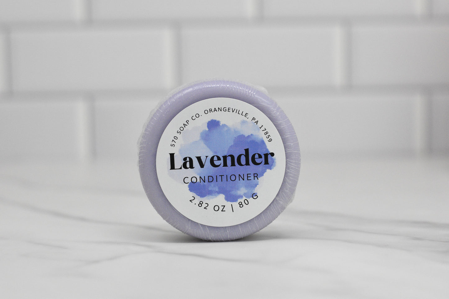 The Lavender Essential Oil Conditioner Bar for All Hair Types by 570 Soap Co. is a round, eco-friendly hair nourishment solution weighing 2.82 oz (80g), and comes in calming purple packaging. It is beautifully displayed on a white marble surface against a tiled wall, showcasing sustainable elegance from Orangeville, PA.