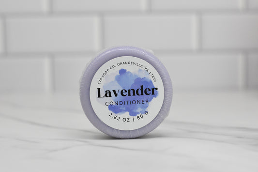 The Lavender Essential Oil Conditioner Bar for All Hair Types by 570 Soap Co. is a round, eco-friendly hair nourishment solution weighing 2.82 oz (80g), and comes in calming purple packaging. It is beautifully displayed on a white marble surface against a tiled wall, showcasing sustainable elegance from Orangeville, PA.