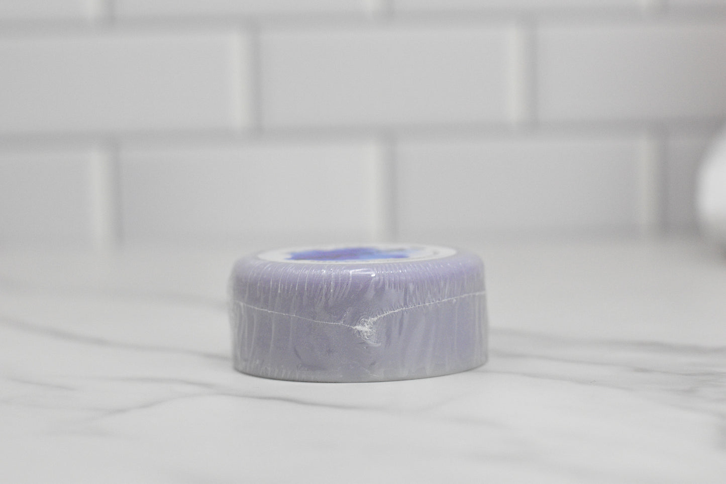 A circular purple soap bar resembling the Conditioner Bar for All Hair Types - Lavender Essential Oil by 570 Soap Co. is wrapped in plastic and sits on a marble surface with a blurred white tiled wall in the background, adding an element of eco-friendly hair care.