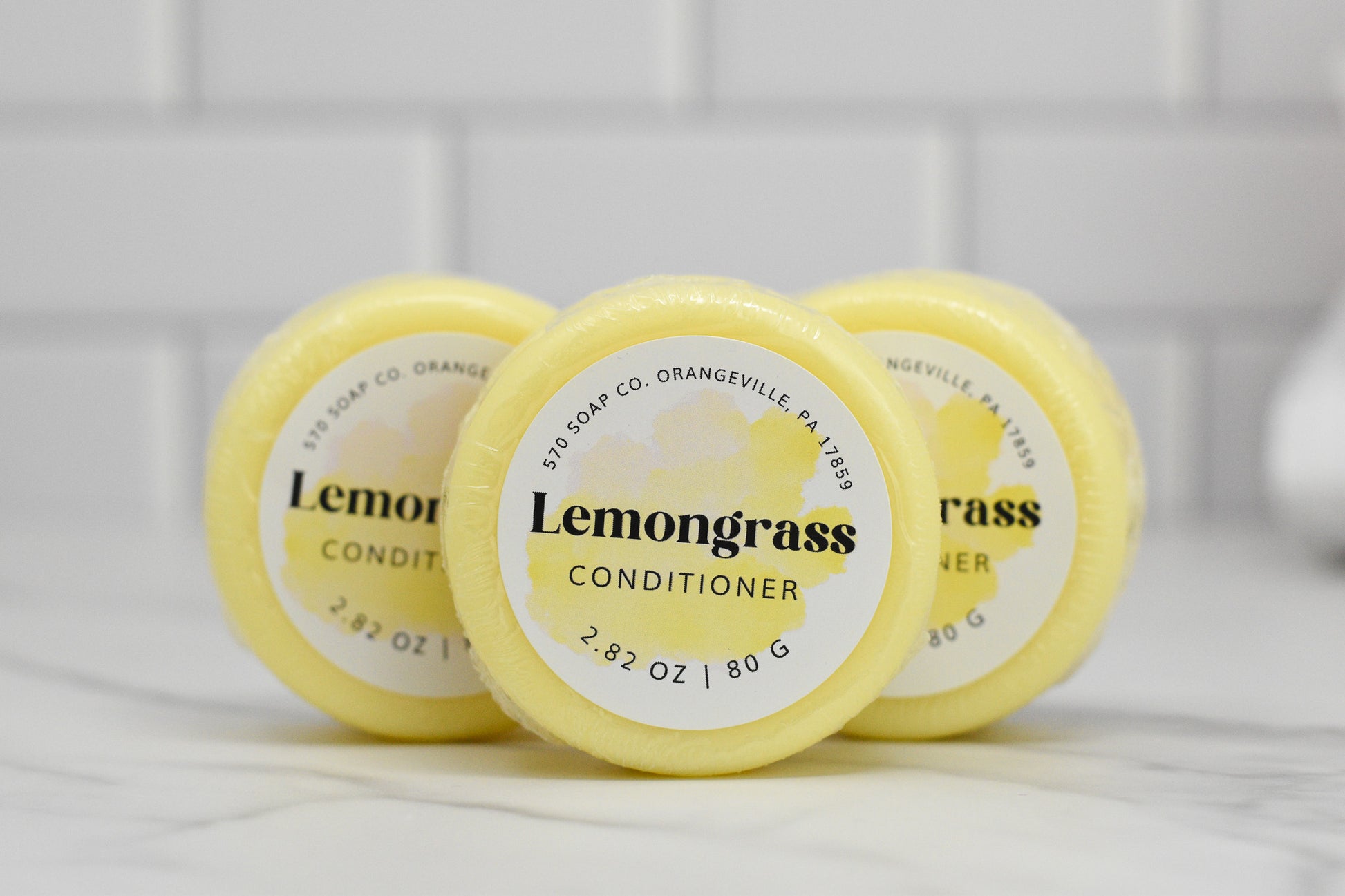 Three round yellow bars of the 570 Soap Co. Conditioner Bar for All Hair Types - Lemongrass Essential Oil stand upright on a white marble surface, against a blurred white tiled background. Each eco-friendly hair care bar weighs 2.82 oz (80 g), promising a moisturizing conditioner experience.