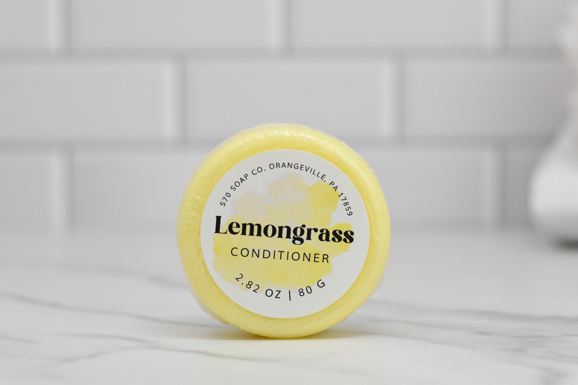 A circular yellow Conditioner Bar for All Hair Types - Lemongrass Essential Oil rests on a white marble surface. The label reads 570 Soap Co., Orangeville, PA with the weight 2.82 oz / 80 g. Offering an eco-friendly hair care solution, this moisturizing conditioner is framed by a backdrop of pristine white tile.