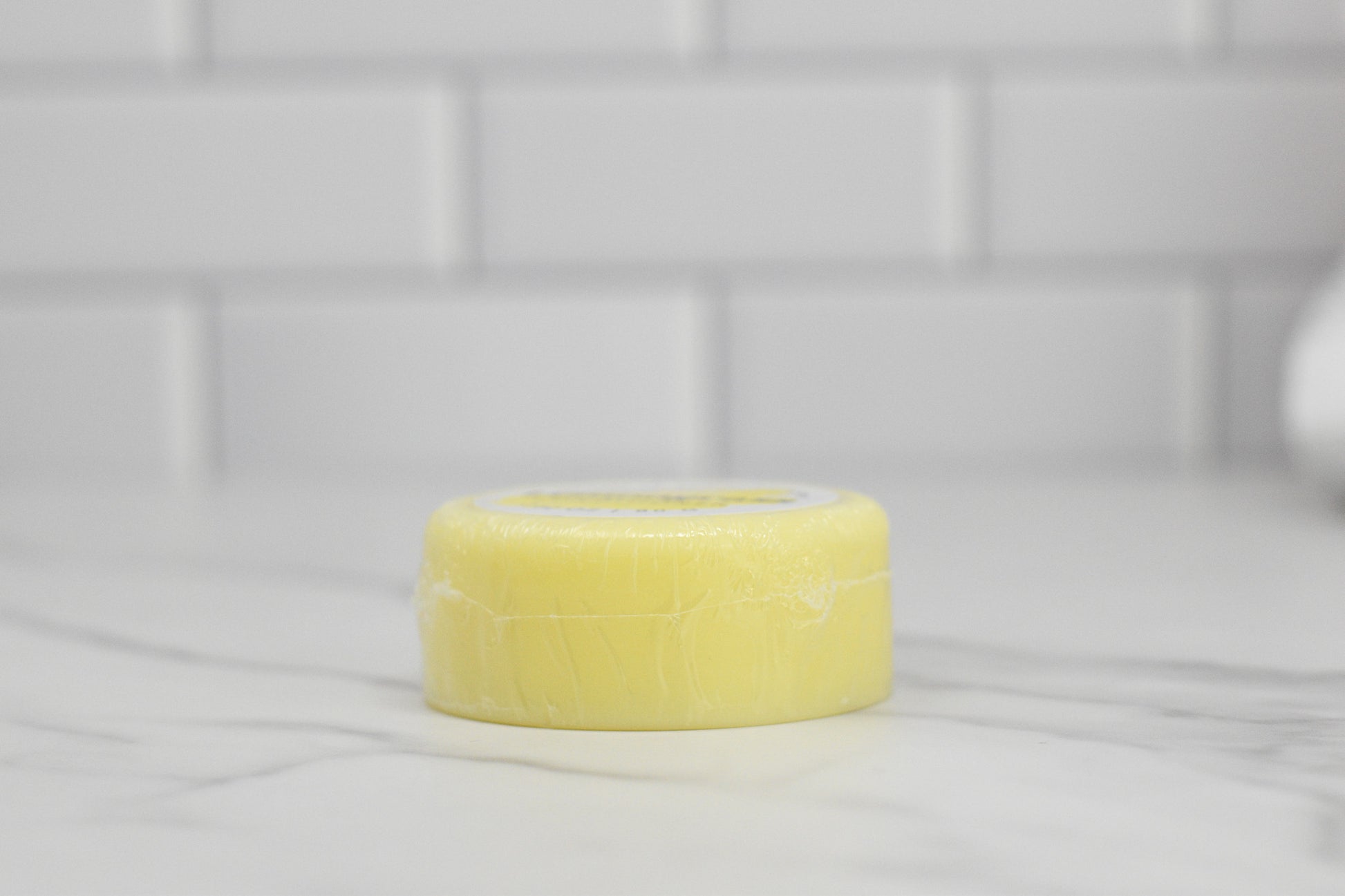 A round, yellow Conditioner Bar for All Hair Types with Lemongrass Essential Oil by 570 Soap Co. sits gracefully on a marble countertop against a backdrop of white subway tiles. Perfect for kitchen or bathroom settings, this eco-friendly hair care product provides a touch of luxury while serving as a moisturizing conditioner.