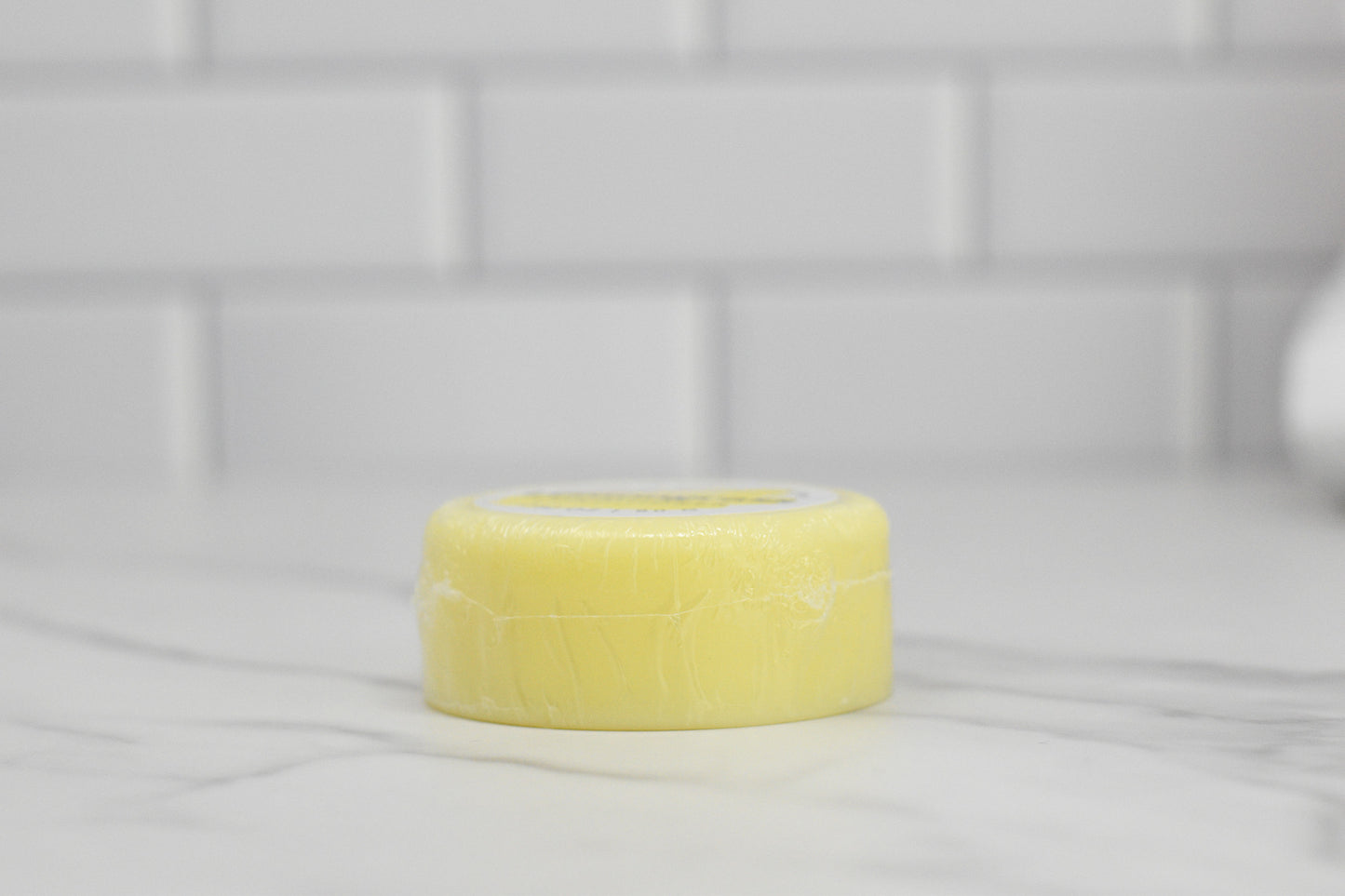 A round, yellow Conditioner Bar for All Hair Types with Lemongrass Essential Oil by 570 Soap Co. sits gracefully on a marble countertop against a backdrop of white subway tiles. Perfect for kitchen or bathroom settings, this eco-friendly hair care product provides a touch of luxury while serving as a moisturizing conditioner.