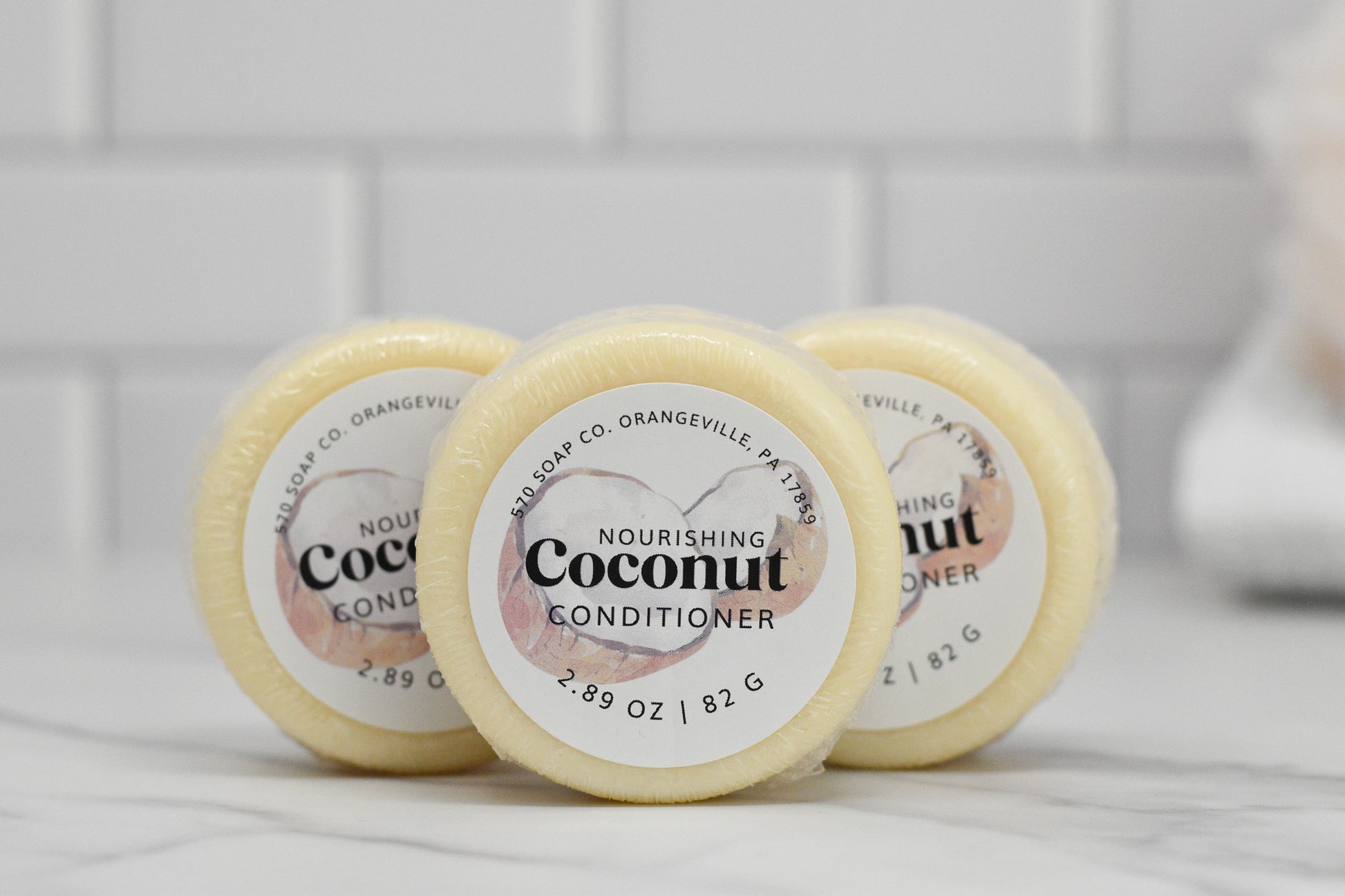 Three round bars of the Nourishing Conditioner Bar for Extra Moisture - Coconut, weighing 2.89 oz / 82 g each and crafted by 570 Soap Co. in Orangeville, are neatly arranged in a row. The white tiled wall in the background emphasizes their gentle care for your hair with botanical extracts.