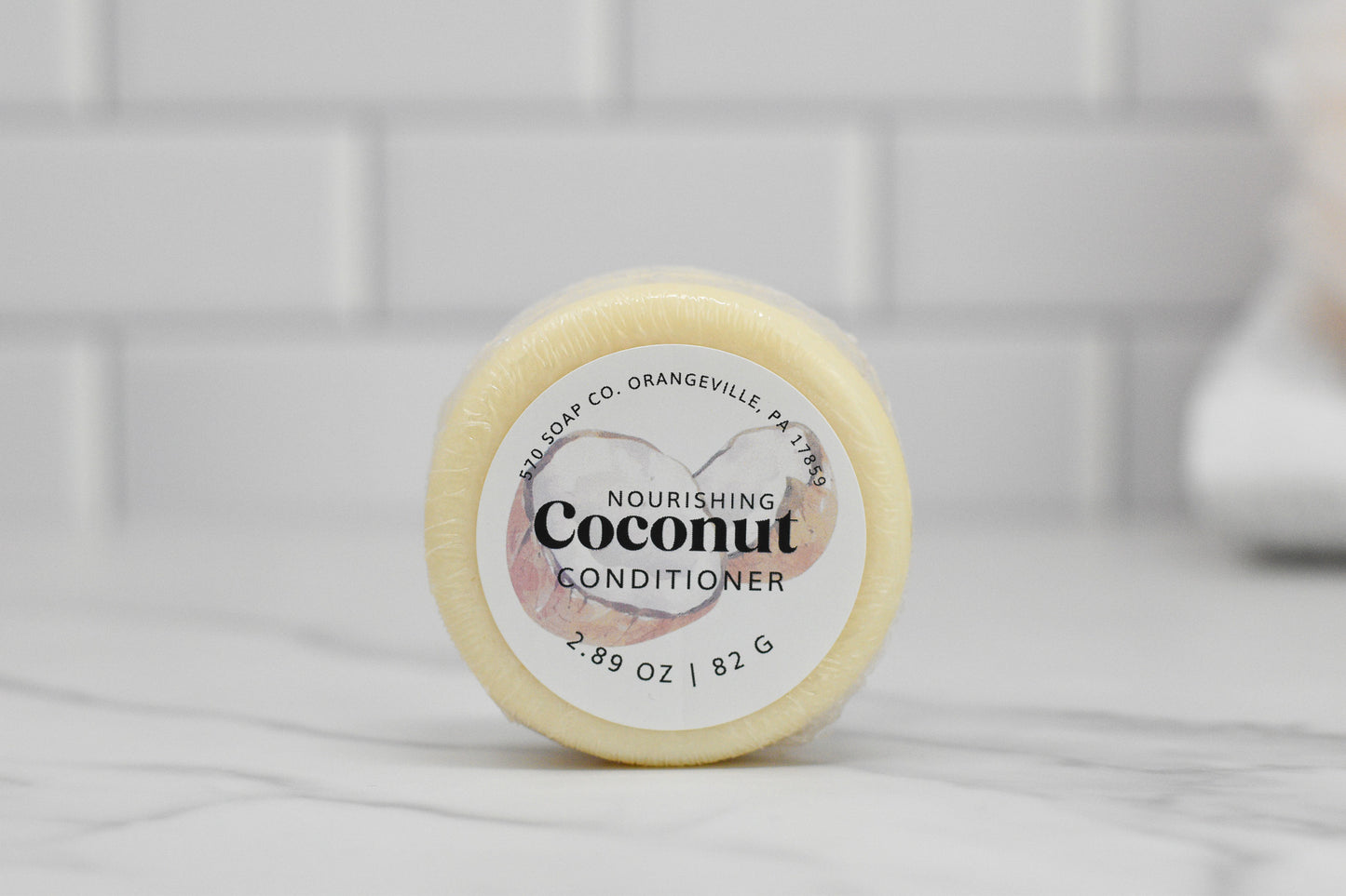 A close-up of the round solid conditioner bar labeled "Nourishing Conditioner Bar for Extra Moisture - Coconut" from 570 Soap Co., featuring botanical extracts for nourishing hair care, rests on a marble surface against a white tile wall. It weighs 2.89 oz (82 g).