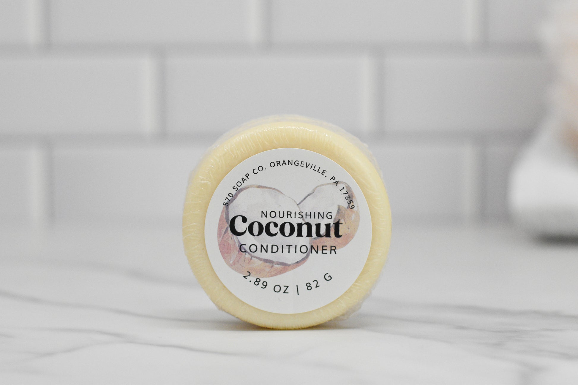 A close-up of the round solid conditioner bar labeled "Nourishing Conditioner Bar for Extra Moisture - Coconut" from 570 Soap Co., featuring botanical extracts for nourishing hair care, rests on a marble surface against a white tile wall. It weighs 2.89 oz (82 g).