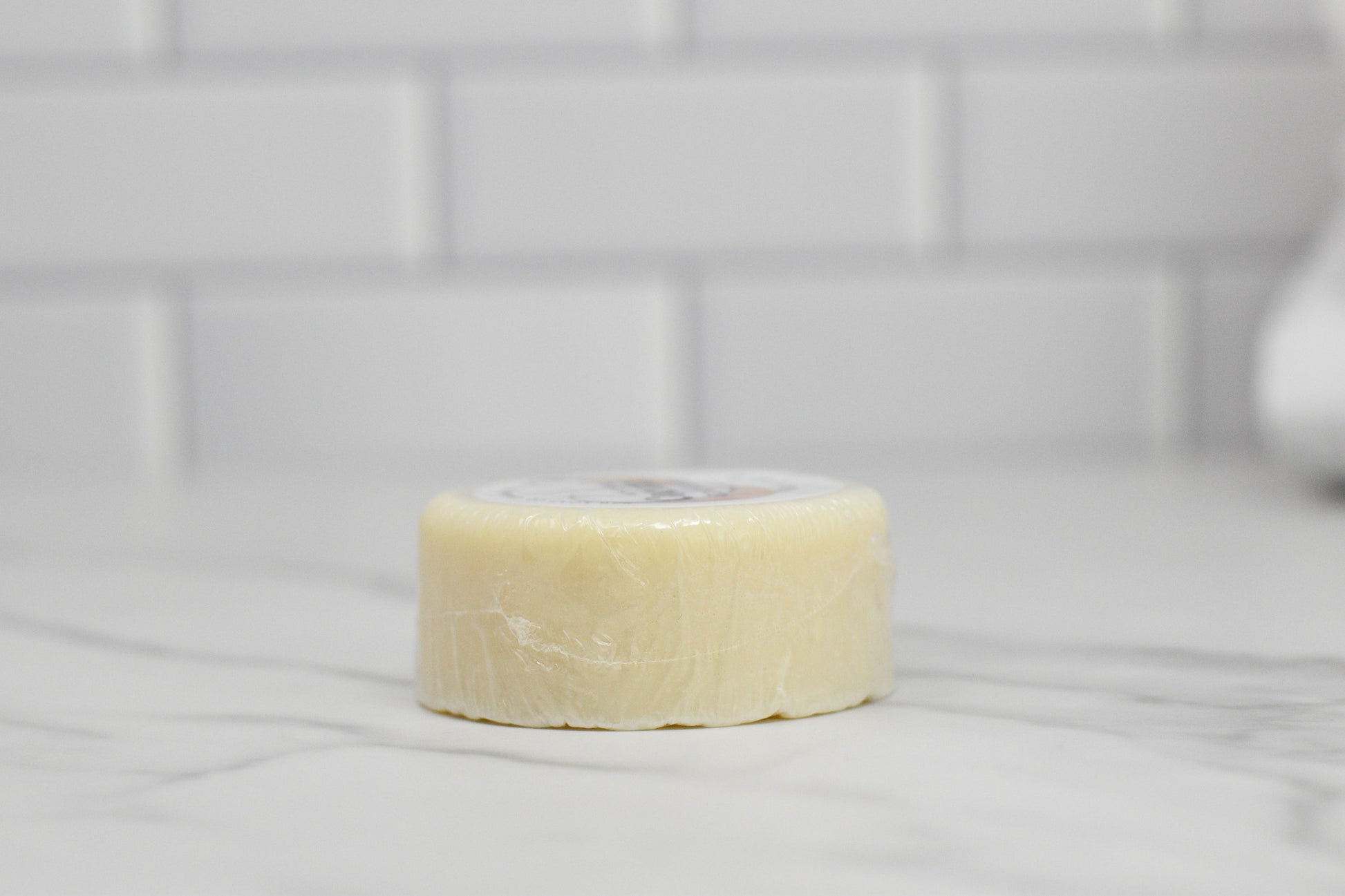 A round bar of 570 Soap Co.'s Nourishing Conditioner Bar for Extra Moisture - Coconut, infused with botanical extracts, rests on a marble countertop wrapped in clear plastic. A white subway tile backsplash highlights its minimalist appeal, with the packaging subtly visible from the top, suggesting its nourishing hair care benefits.