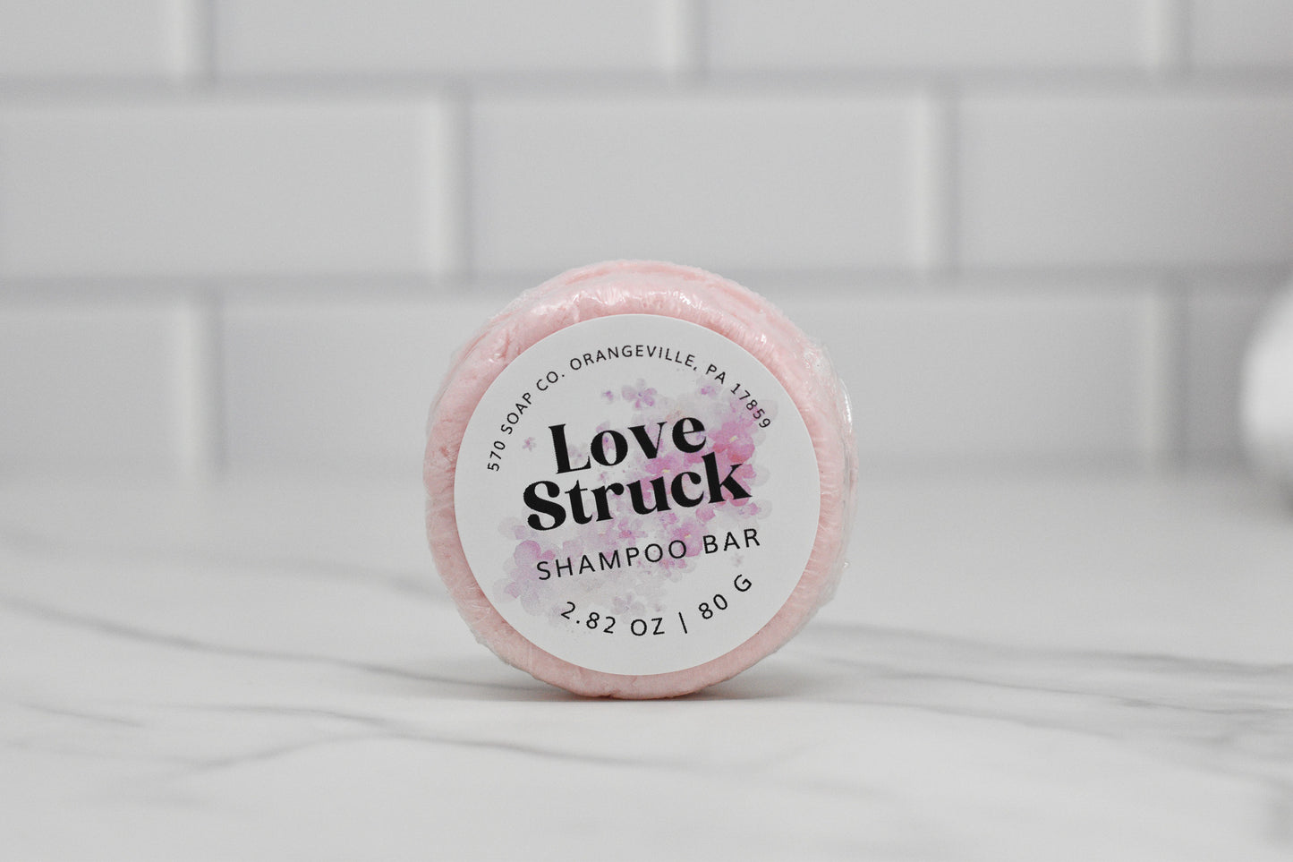 A round, pink Love Struck Solid Shampoo Bar from 570 Soap Co. sits gracefully on a marble surface. The label states its weight as 2.82 oz (80 g). With white subway tiles in the background, this eco-friendly shampoo bar is perfect for a sustainable beauty routine.