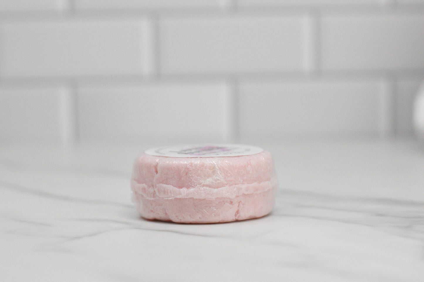 A round, pale pink Love Struck Solid Shampoo Bar from 570 Soap Co. sits wrapped in transparent plastic on a white marble countertop. The backdrop showcases a minimalist white tiled wall, beautifully enhancing your eco-friendly shampoo bar for a sustainable beauty routine.