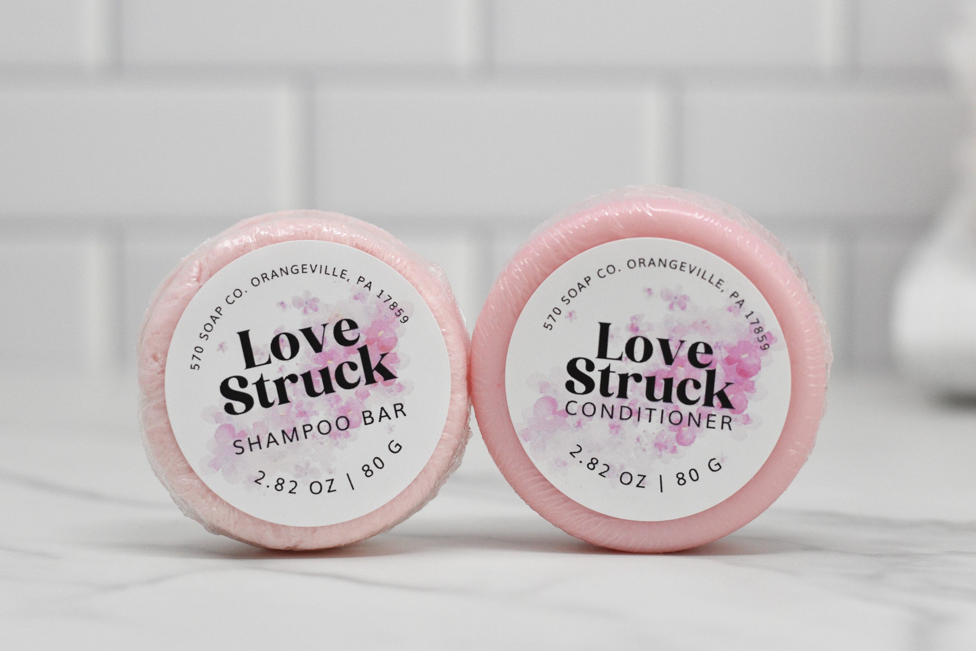 Two round, pink soap bars rest on a marble surface. The left, a luxurious Love Struck Shampoo Bar, and the right, an eco-friendly Conditioner Bar for All Hair Types - Love Struck. Both have a floral design and are crafted by 570 Soap Co. in Orangeville, PA.