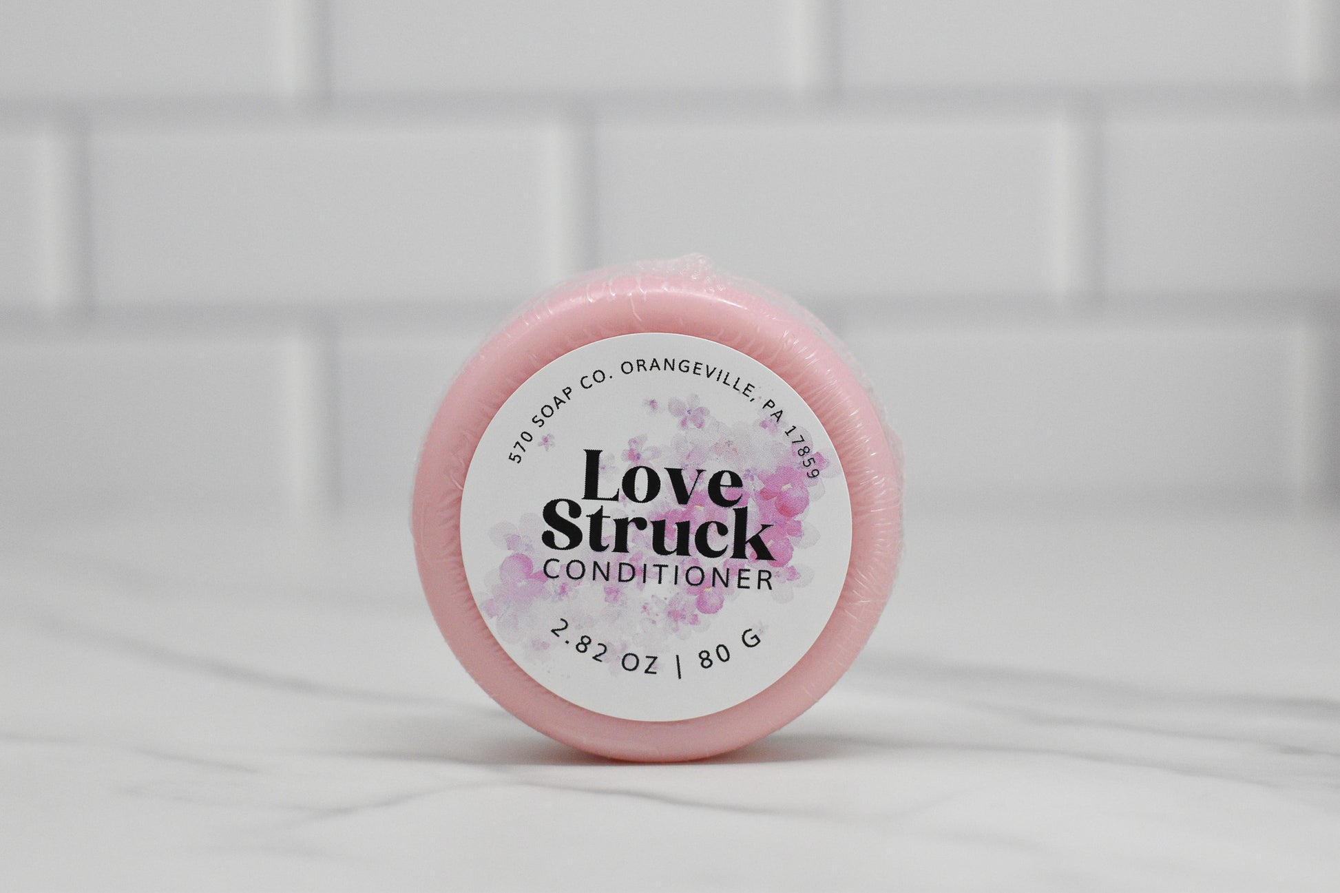 A round pink container of 570 Soap Co.'s Conditioner Bar for All Hair Types - Love Struck sits elegantly on a marble surface. The label, adorned with floral designs and weight details (2.82 oz / 80 g), highlights this eco-friendly alternative in luxurious hair care against a backdrop of white subway tiles.