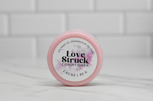 A round pink container of 570 Soap Co.'s Conditioner Bar for All Hair Types - Love Struck sits elegantly on a marble surface. The label, adorned with floral designs and weight details (2.82 oz / 80 g), highlights this eco-friendly alternative in luxurious hair care against a backdrop of white subway tiles.