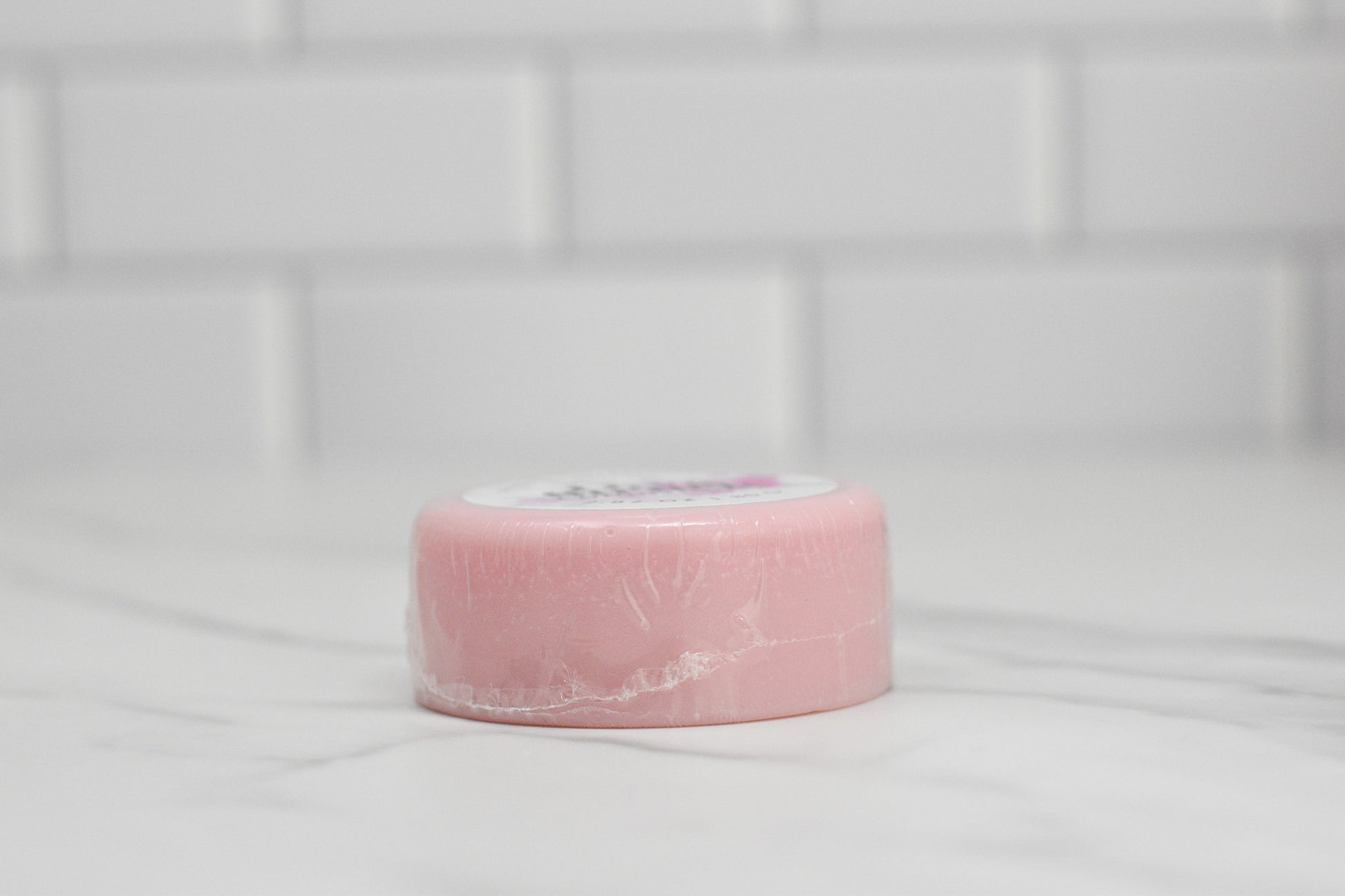 A pink, round Love Struck conditioner bar from 570 Soap Co., encased in clear packaging, sits elegantly on a white marble surface. In the background, the glossy finish of white rectangular tiles highlights this luxurious hair care option as an eco-friendly alternative to traditional bottled conditioners.