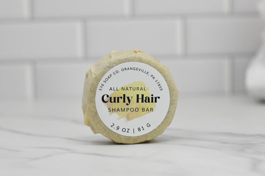 A round, beige Curly Hair Shampoo Bar - All Natural from 570 Soap Co. in Orangeville, PA 17859, weighing 2.9 oz (81 g), rests elegantly on a marble surface against a white tiled background.