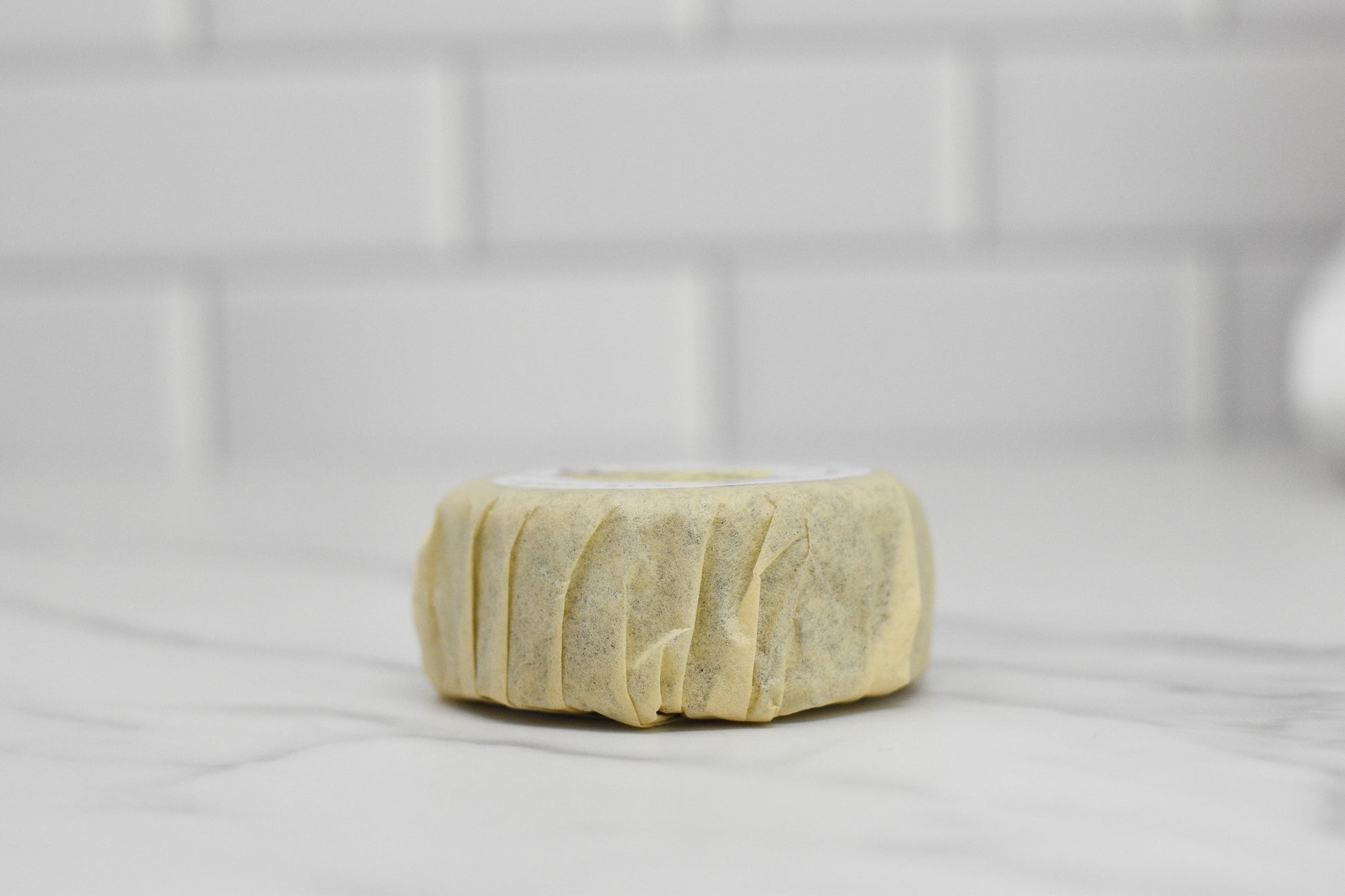 A wrapped round item, the Curly Hair Shampoo Bar from 570 Soap Co., rests on a white marble surface with a white tiled background. The beige paper wrapping adds a rustic charm, perfect for those seeking frizz-free solutions for curly hair.