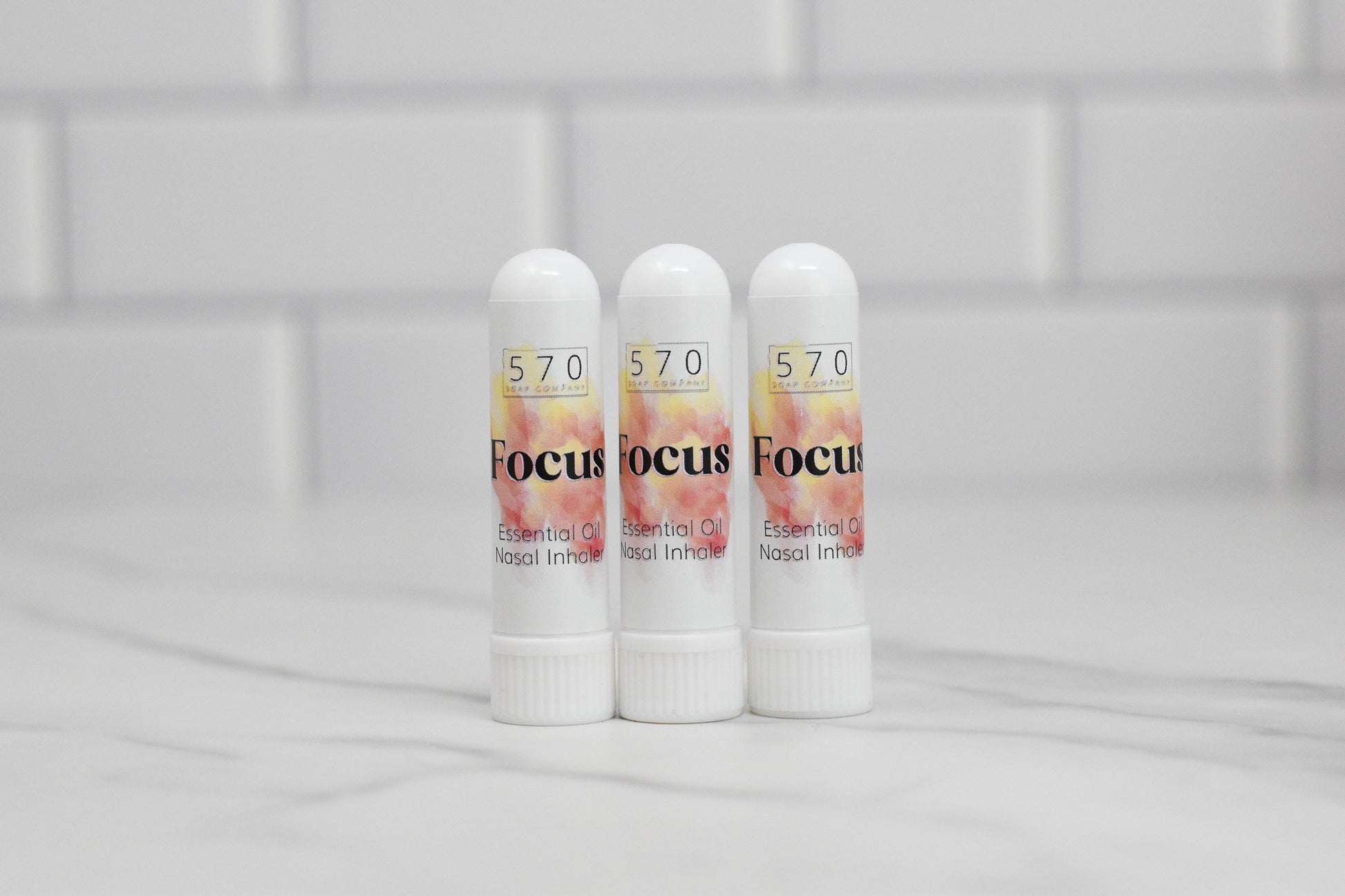 Three white cylindrical nasal inhalers from 570 Soap Co., labeled with "Focus," are arranged on a marble surface against a backdrop of white tiles, providing natural relief through essential oils.